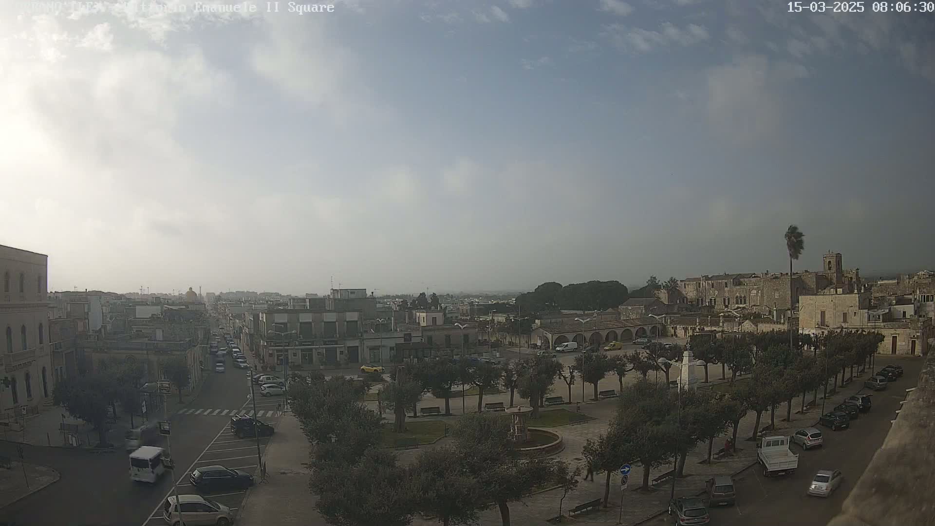 A hazy, partly sunny day overlooks a town square with a fountain, surrounded by trees and parked cars, with a street leading to a larger town in the distance.