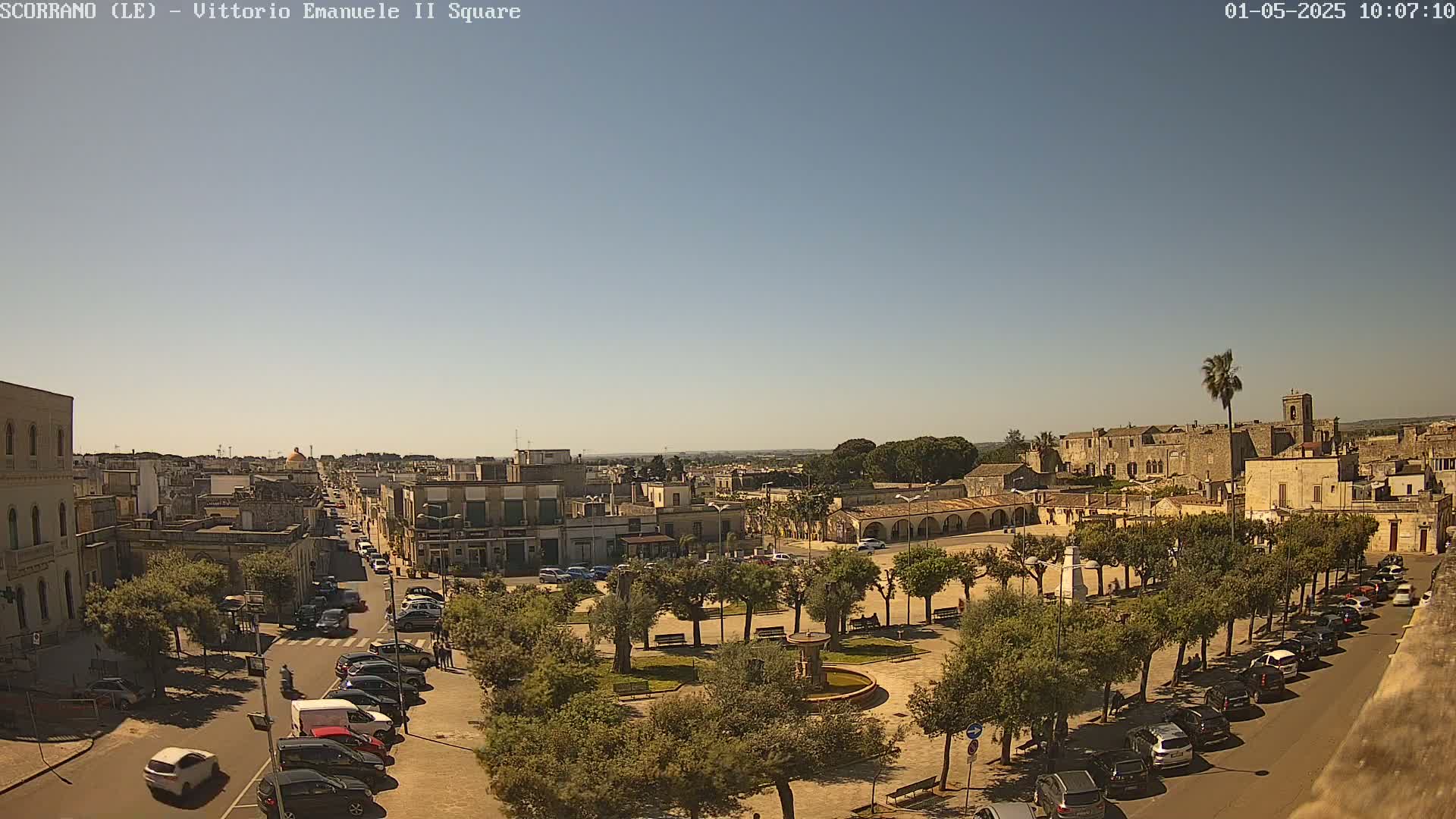 A sunny day reveals a town square with a fountain, surrounded by parked cars and lined with trees, extending to a cityscape under a clear blue sky.