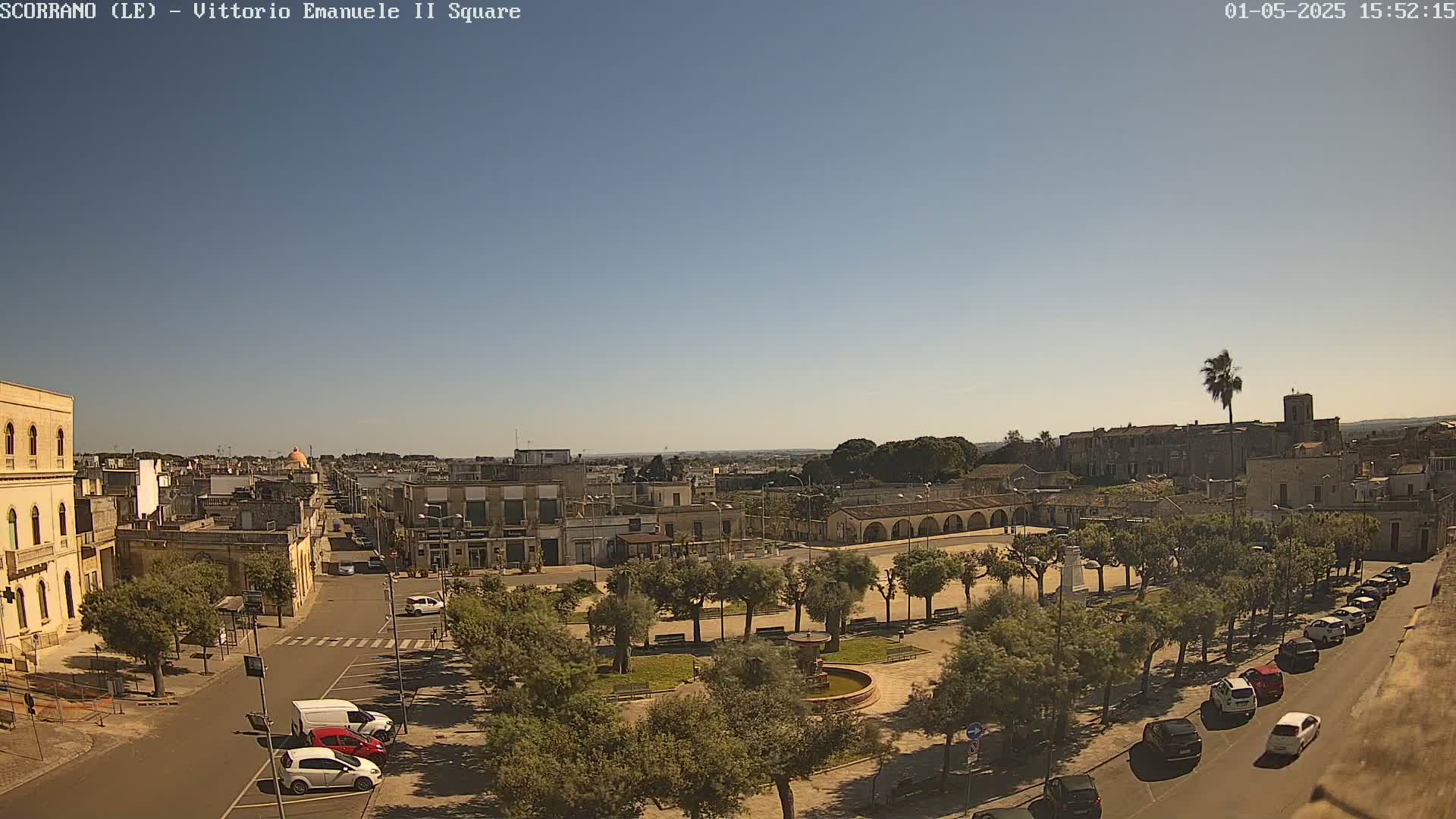 A town square with a central fountain, lined with trees and parked cars, is visible under a clear, sunny sky.