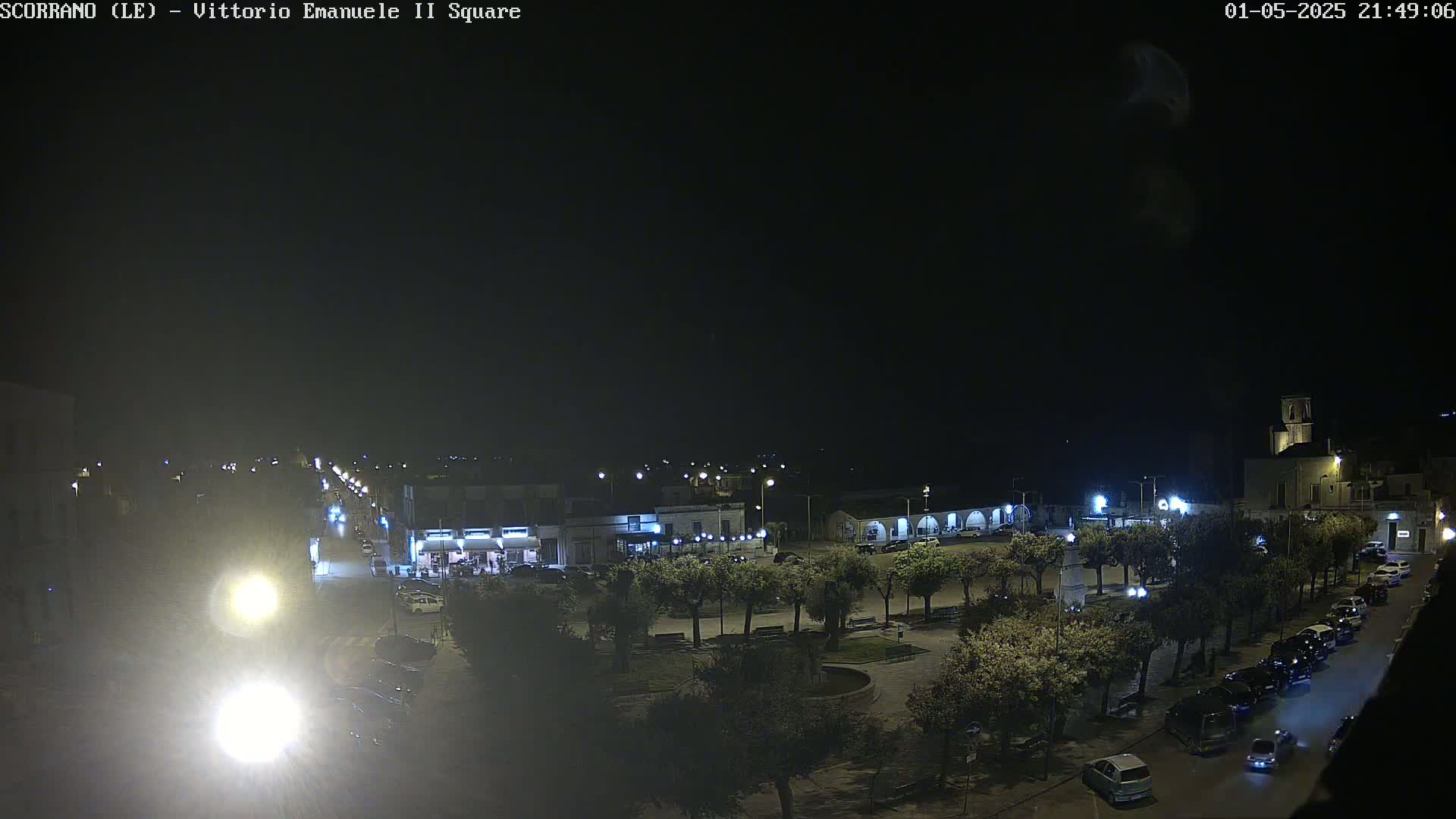 A nighttime view of a town square, illuminated by streetlights, with cars parked along the sides of the streets and trees lining the edges of the square.