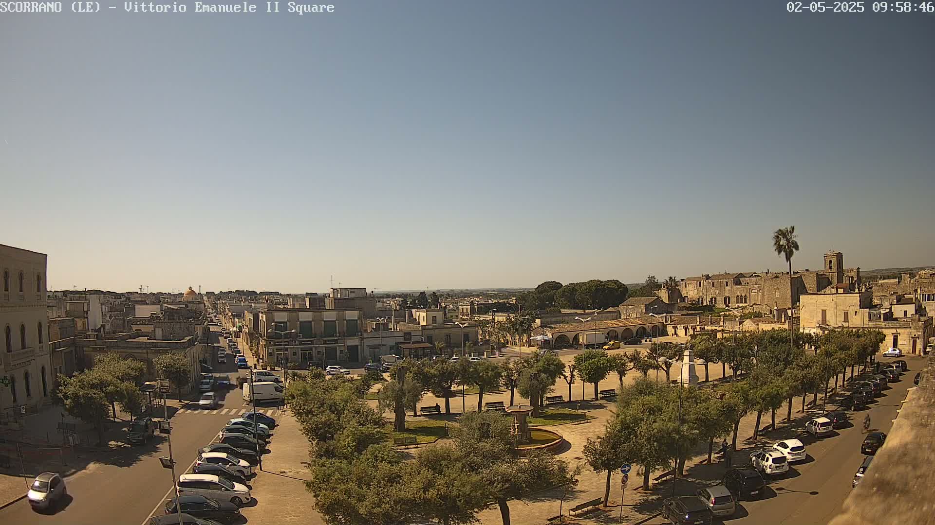 A sunny day reveals a town square with a central fountain, surrounded by trees and parked cars lining the streets, all under a clear blue sky.