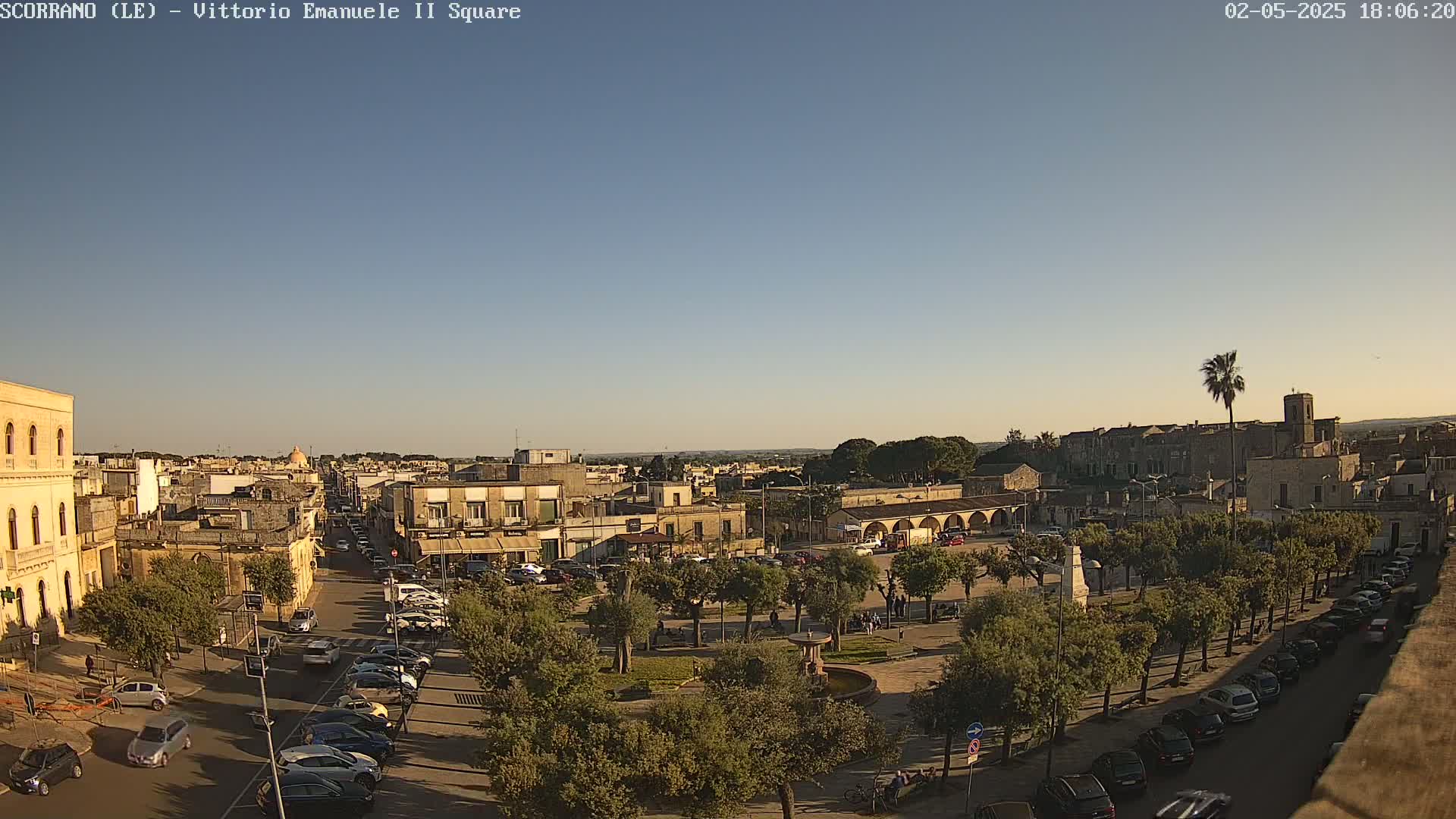 A sunlit town square, surrounded by buildings and trees, with cars parked along the streets under a clear sky.