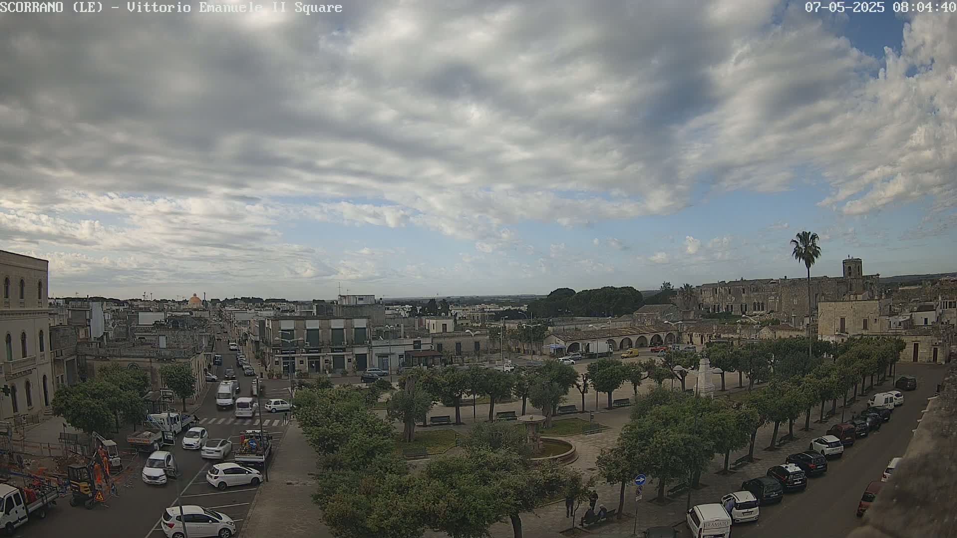 A partly cloudy day reveals a town square with a fountain, surrounded by trees and parked cars, and buildings extending into the distance.