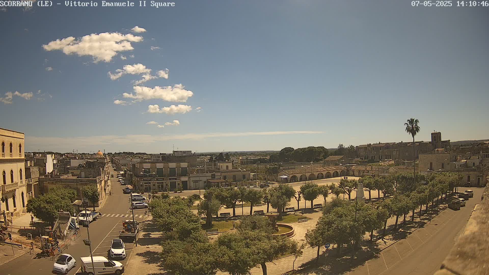A partly sunny day reveals a town square with a fountain, surrounded by trees and buildings, and a street with parked cars leading towards a larger town in the distance.