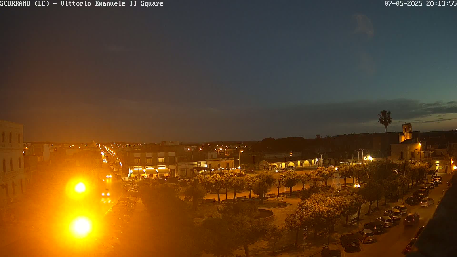 A nighttime view of a town square, illuminated by streetlights, with cars parked along the edges and trees lining the pathways under a mostly clear, dark blue twilight sky.