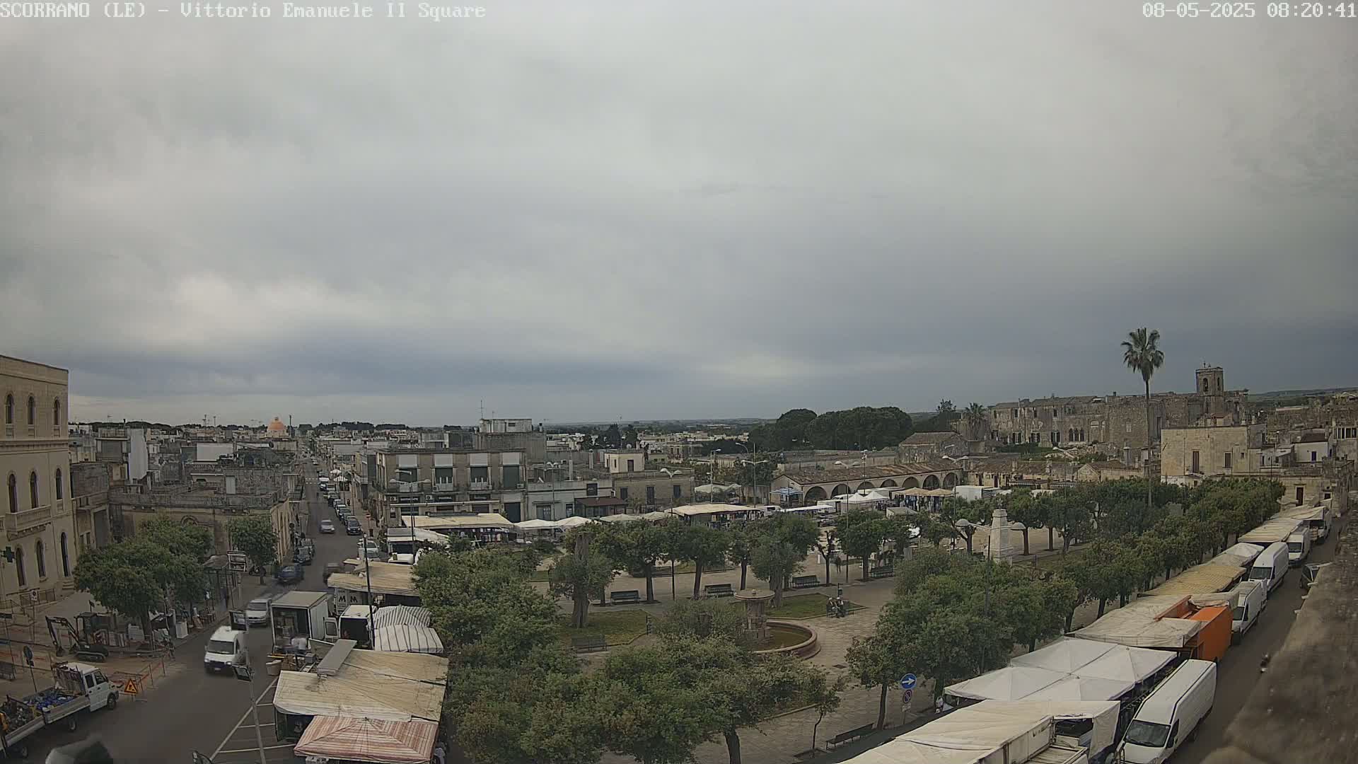 An overcast sky hangs over a town square filled with market stalls and vehicles, with a street leading to the city beyond.