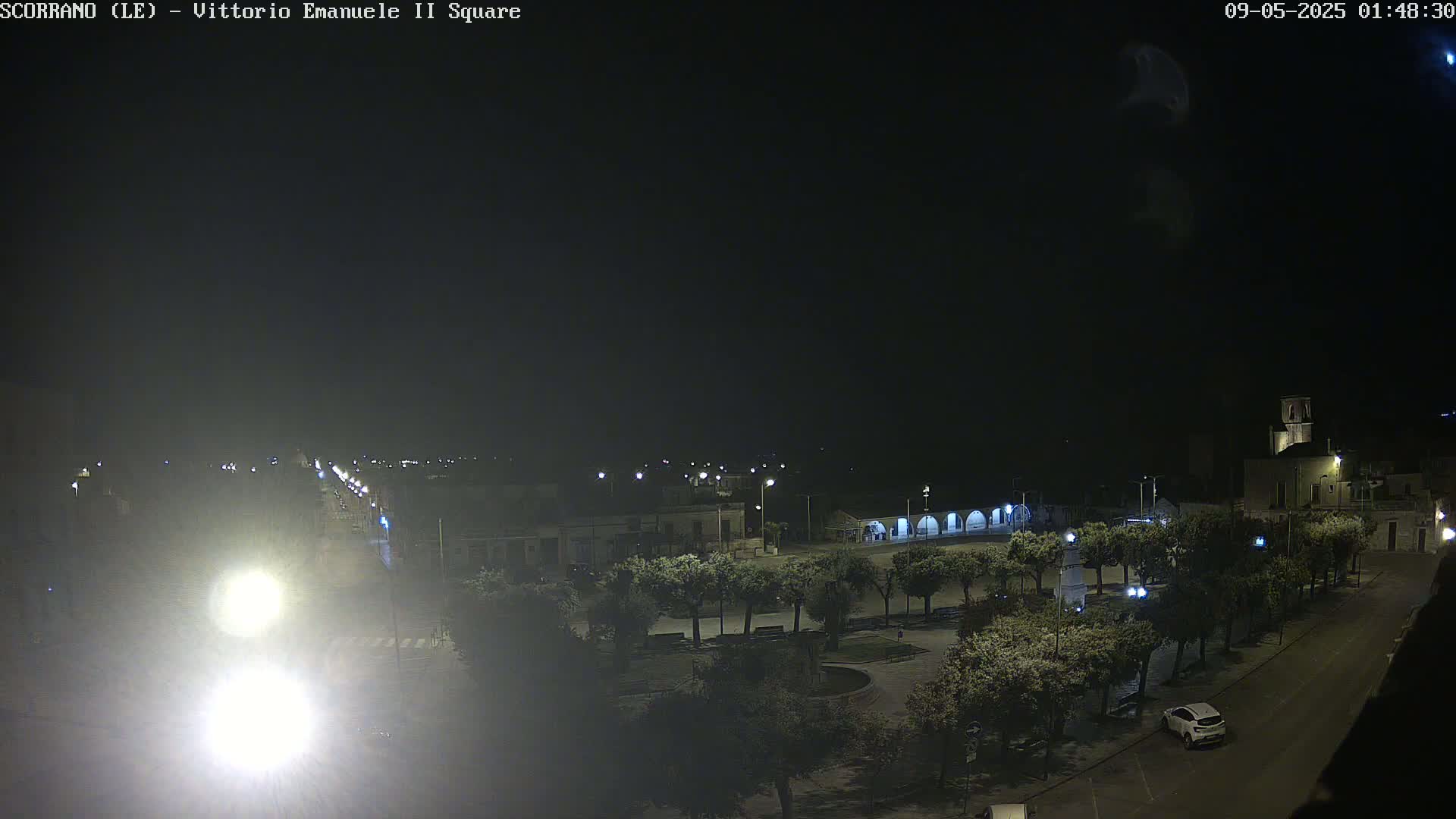 A nighttime, outdoor view of a town square with trees, buildings, streetlights, and a single car parked.
