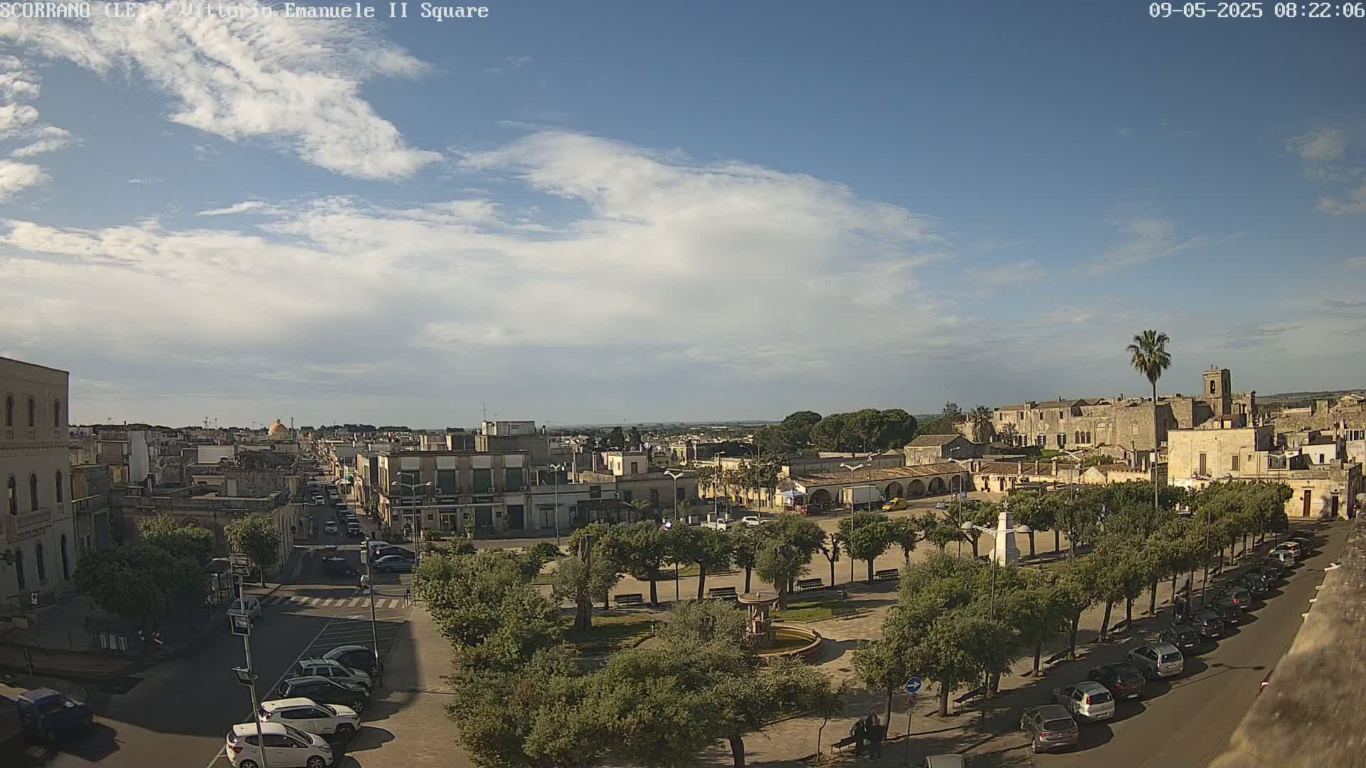 A partly cloudy sky looks down on a town square with a fountain, surrounded by buildings and trees, and cars parked along the streets.