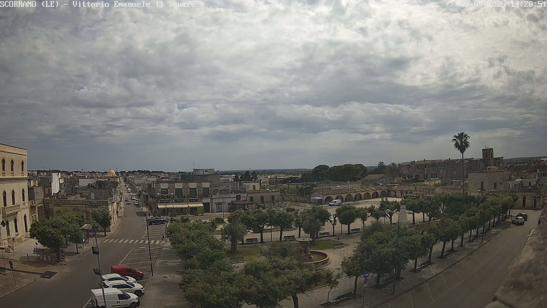 An elevated view shows a town square with a fountain and surrounding trees under a mostly cloudy sky.