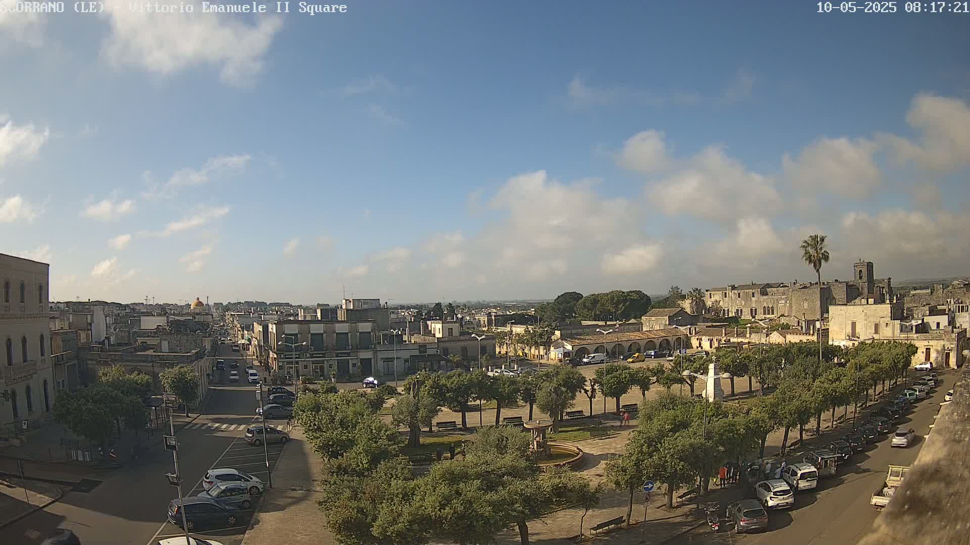 A partly cloudy day reveals a town square with a central fountain, surrounded by trees and parked cars, and a larger town extending into the distance.