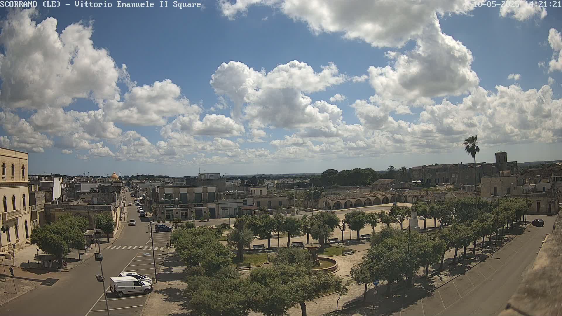 A partly sunny day reveals a town square with a fountain, surrounded by trees and buildings, and a street leading away from it.