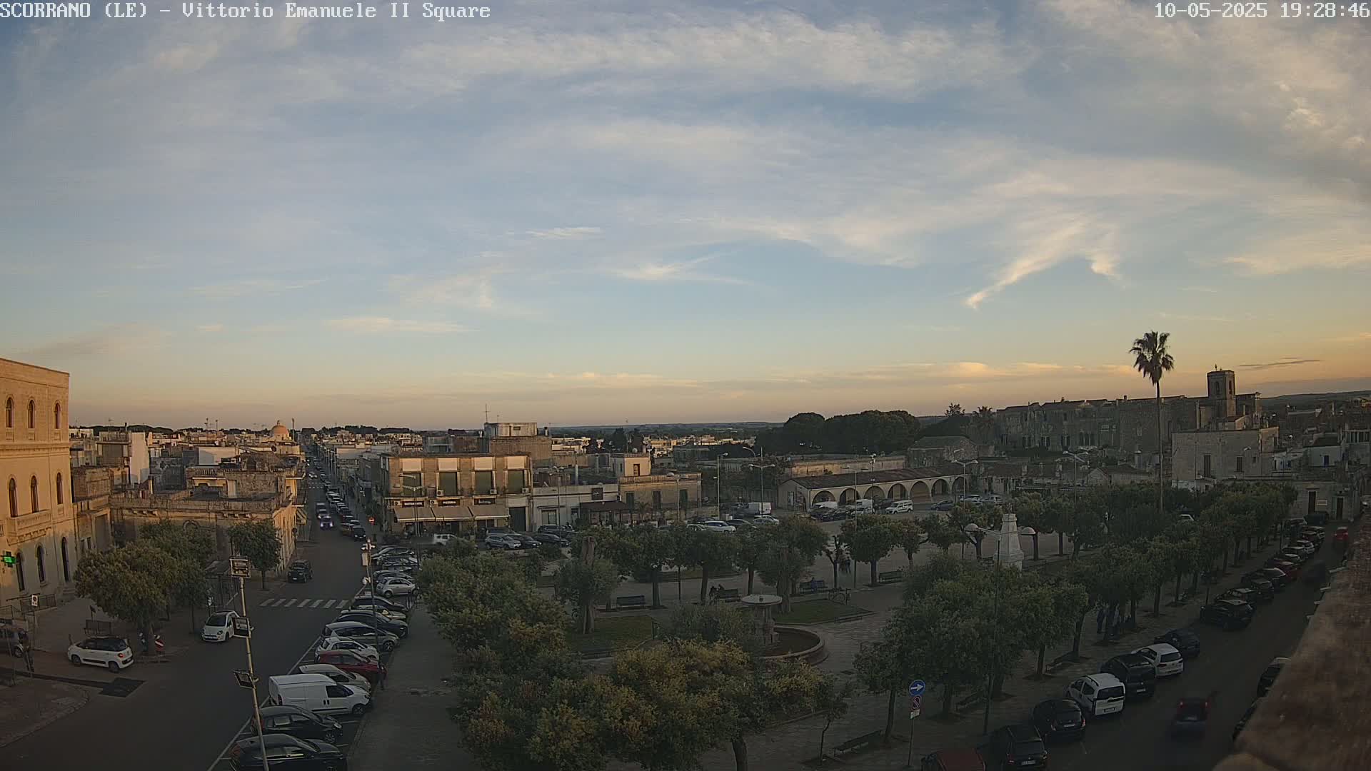 An aerial view of a town square, with cars parked along the streets and trees lining the square, under a partly cloudy sunset sky.