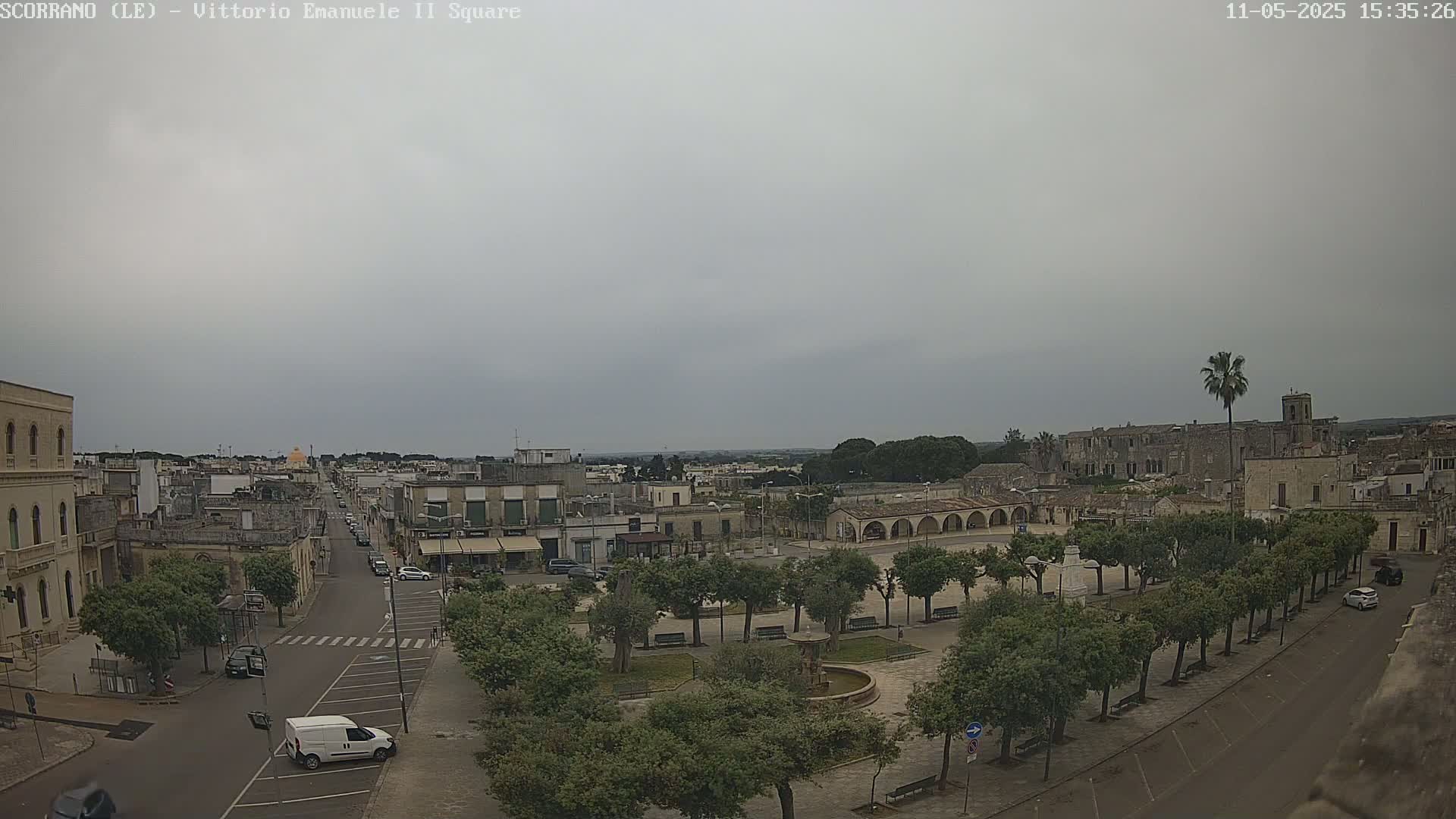 A town square with a fountain and numerous trees is shown under an overcast sky.