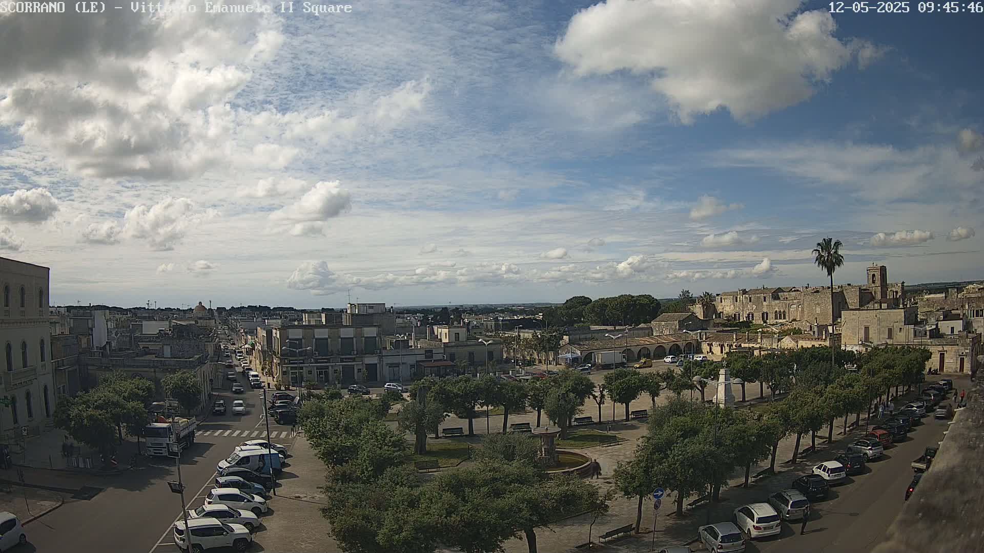 A partly cloudy sky overlooks a town square with parked cars lining the streets, trees lining the square, and buildings surrounding it.