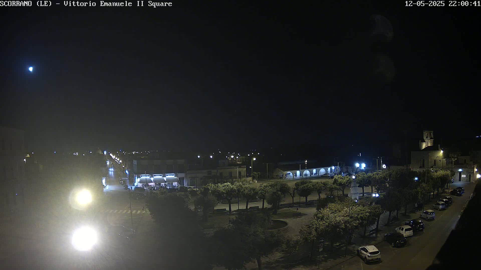 A nighttime, aerial view of a town square with parked cars and trees, under a dark sky with a single visible light source in the distance.