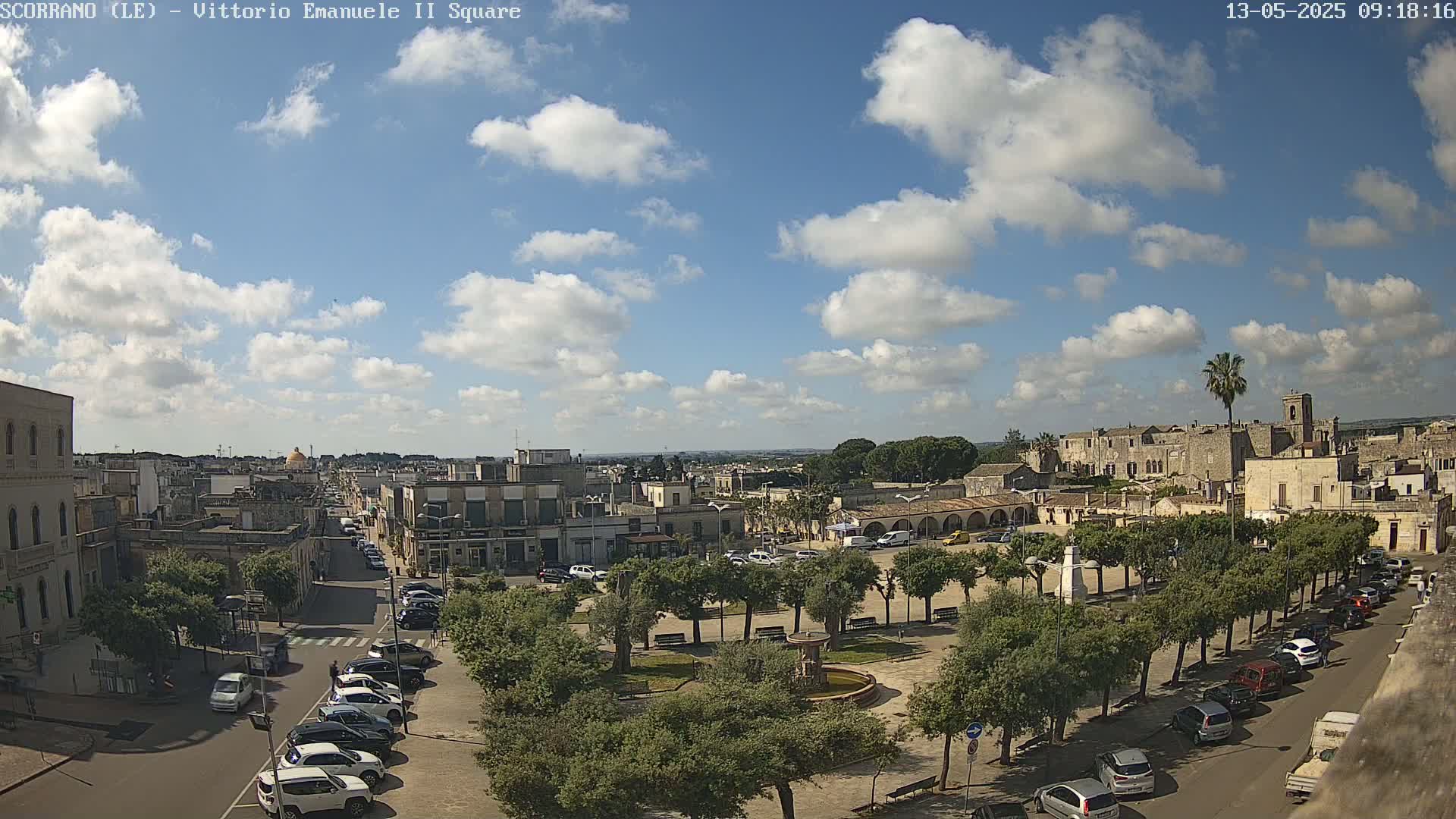 A town square with parked cars and a fountain is surrounded by buildings under a partly cloudy sky.