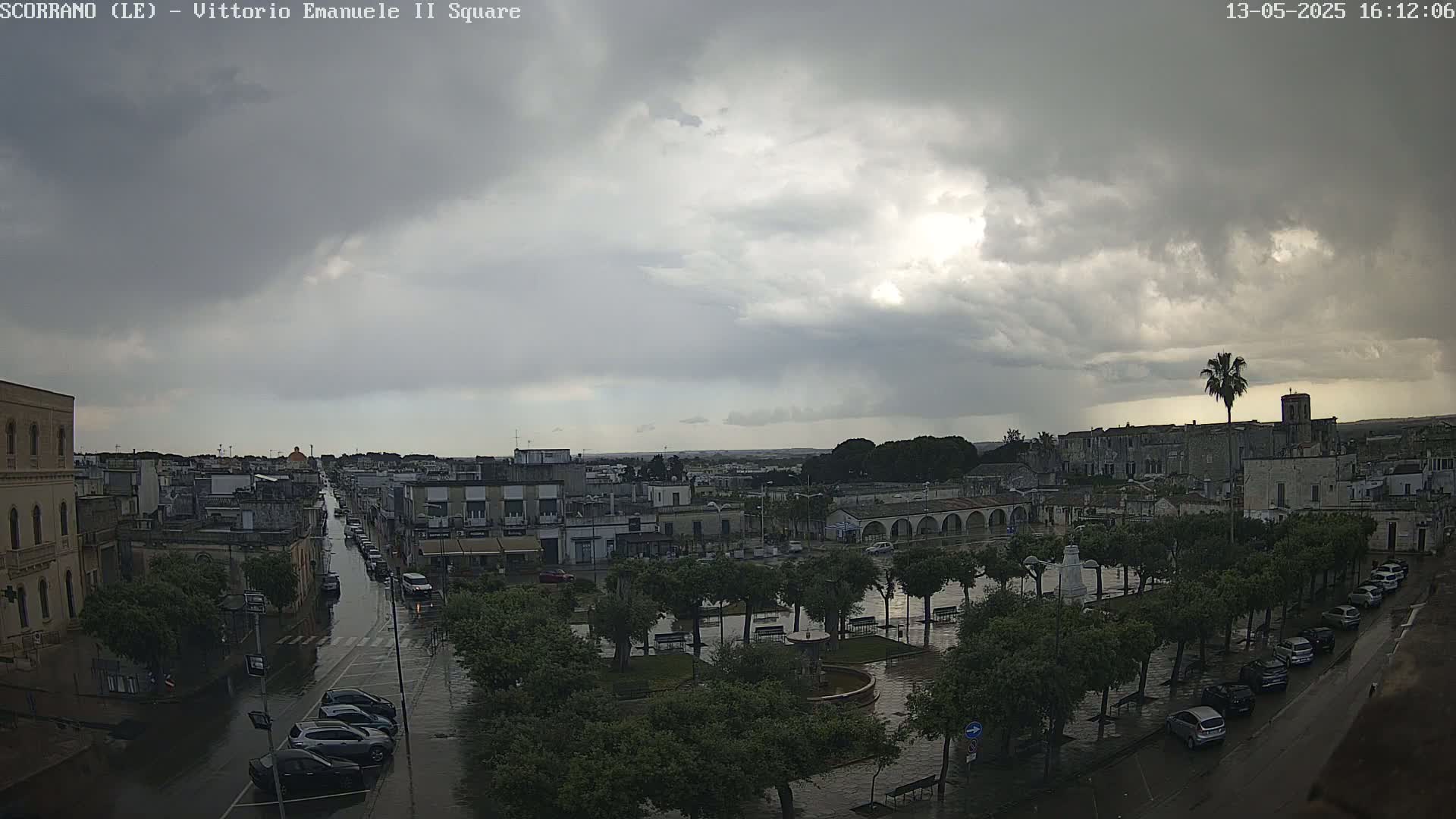A rain-soaked town square and street, under a dark, cloudy sky, are surrounded by buildings and trees.