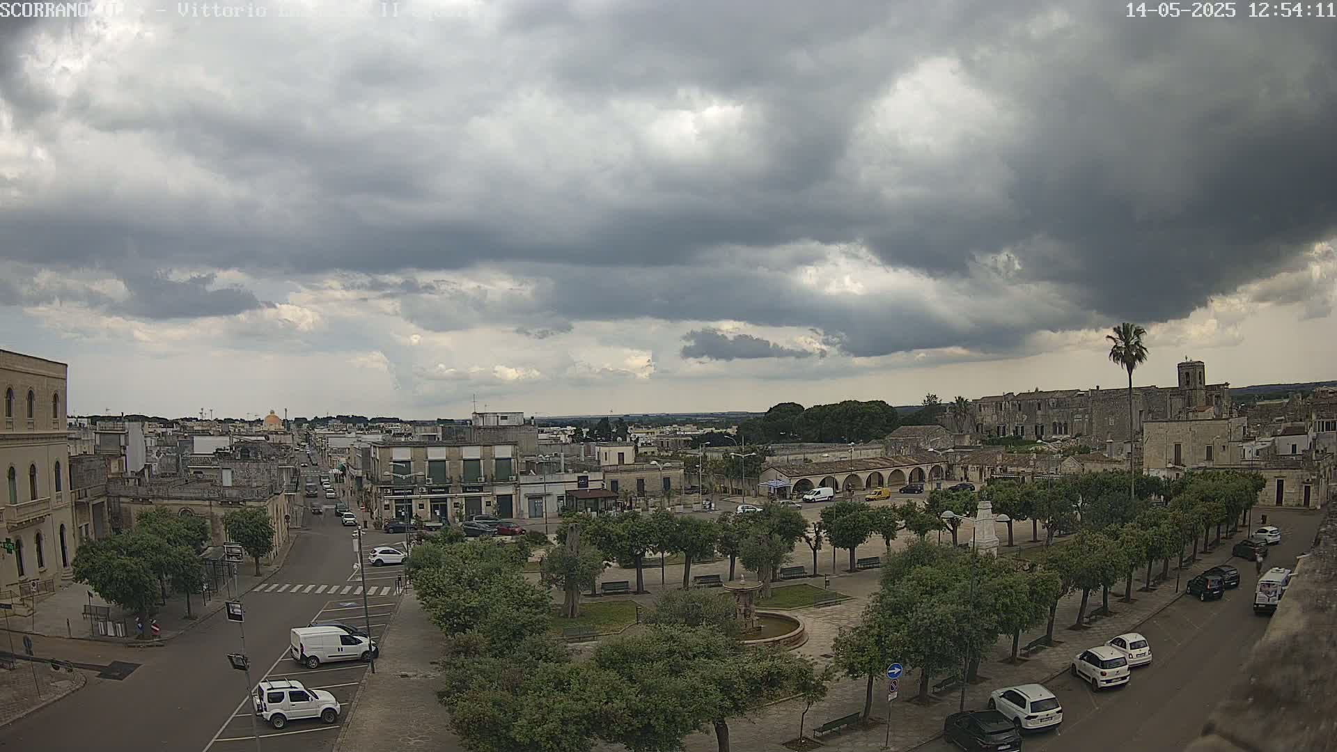 A town square with a fountain and numerous trees is surrounded by buildings under a heavily clouded sky.