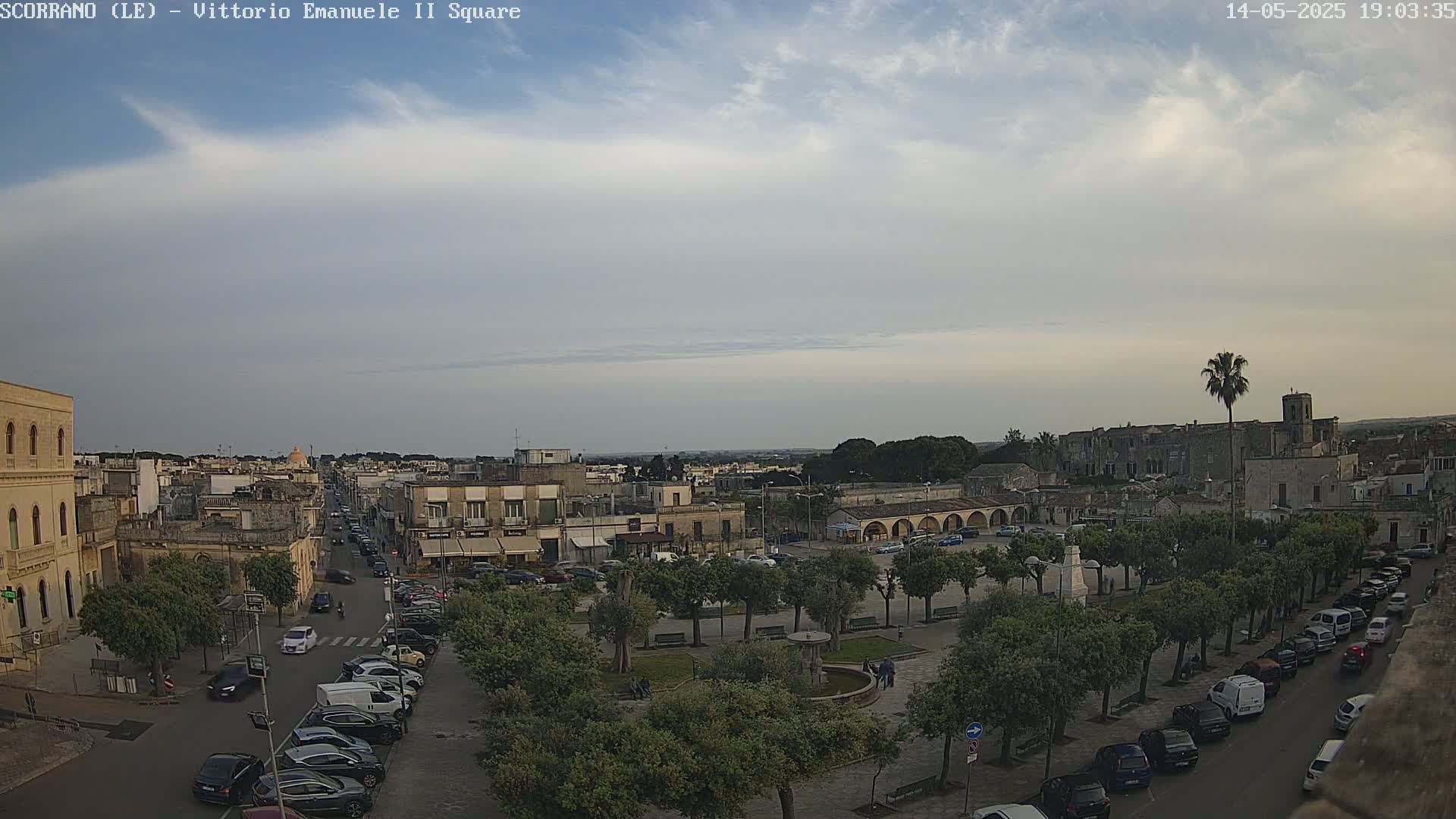 A mostly cloudy day reveals a town square with parked cars, trees, and buildings, extending to a cityscape in the distance.