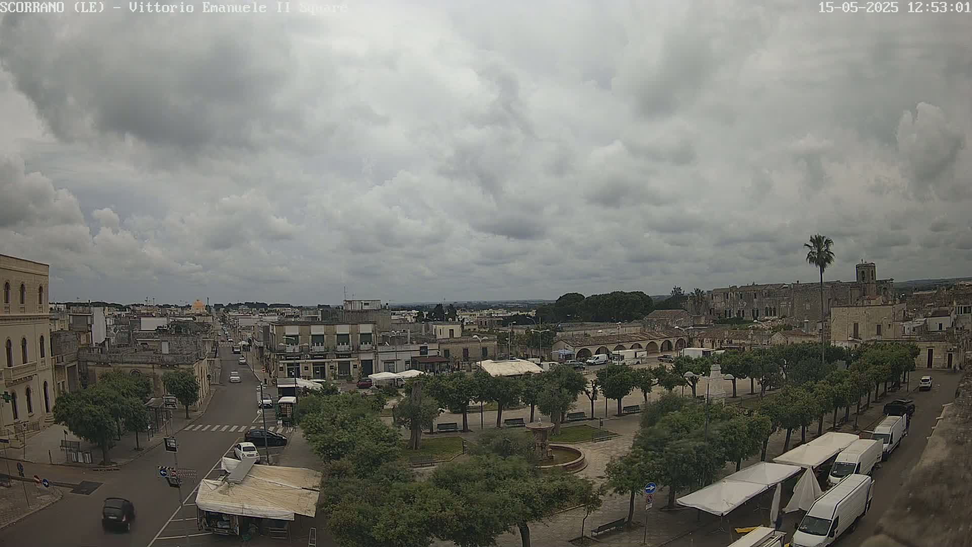 A partly cloudy day overlooks a town square with a fountain, trees, and buildings, including a large structure in the background.
