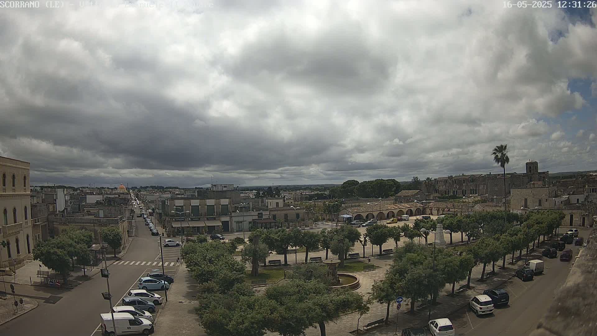 A mostly cloudy sky overlooks a town square with parked cars and a fountain, a street lined with parked cars leads away from the square to a larger town.