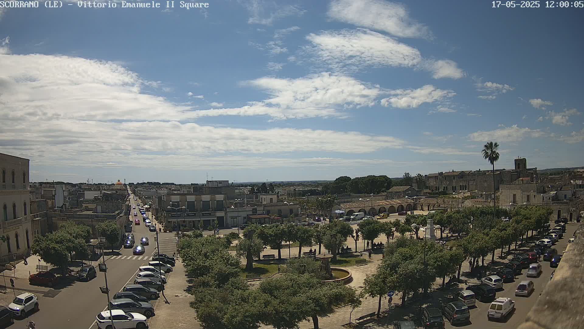 A partly cloudy day overlooks a town square with a central fountain, surrounded by trees and parked cars, with a street leading away from it filled with more vehicles.