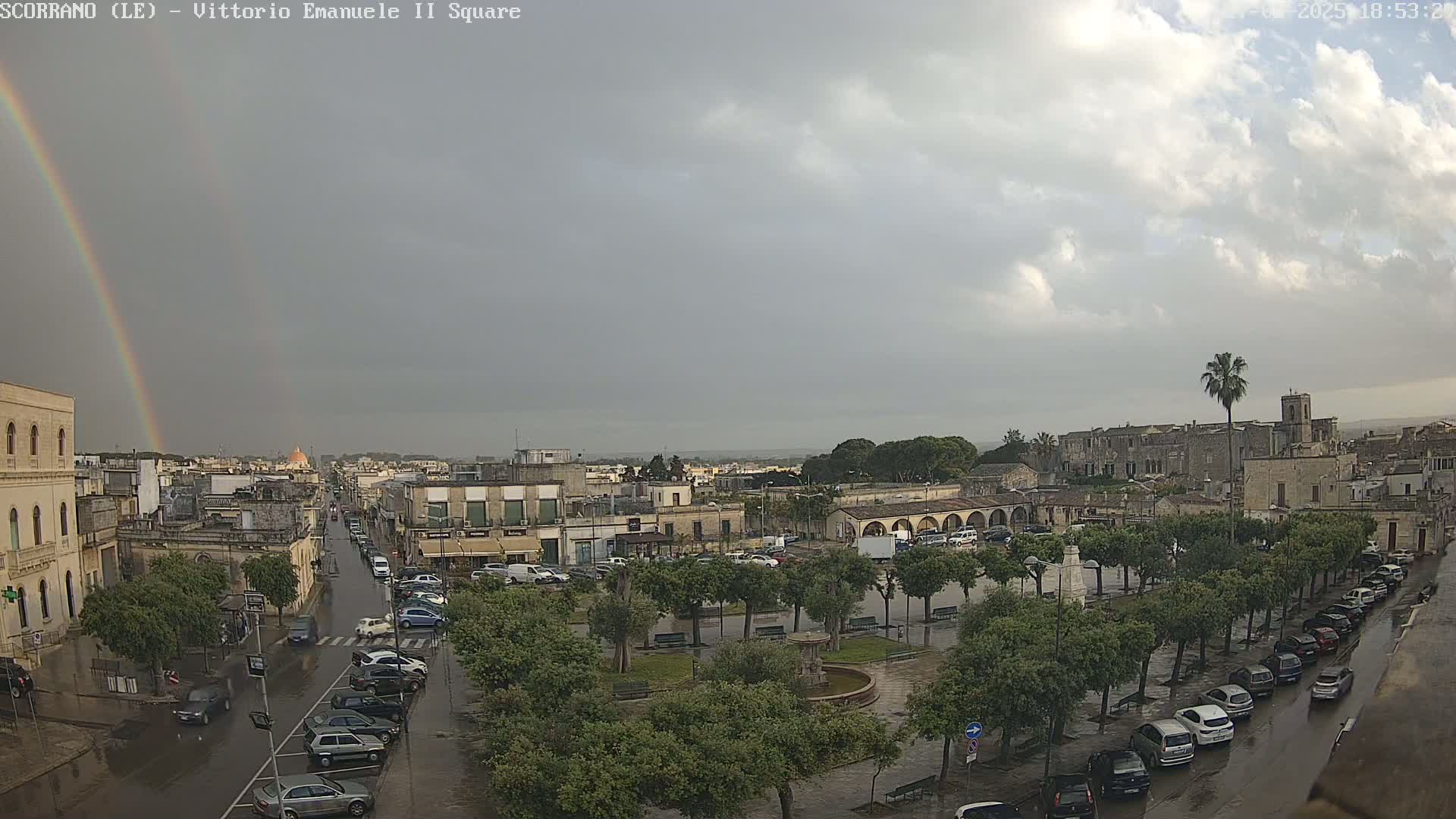 A rain-soaked town square, partially surrounded by trees and parked cars, features a rainbow in the overcast sky.