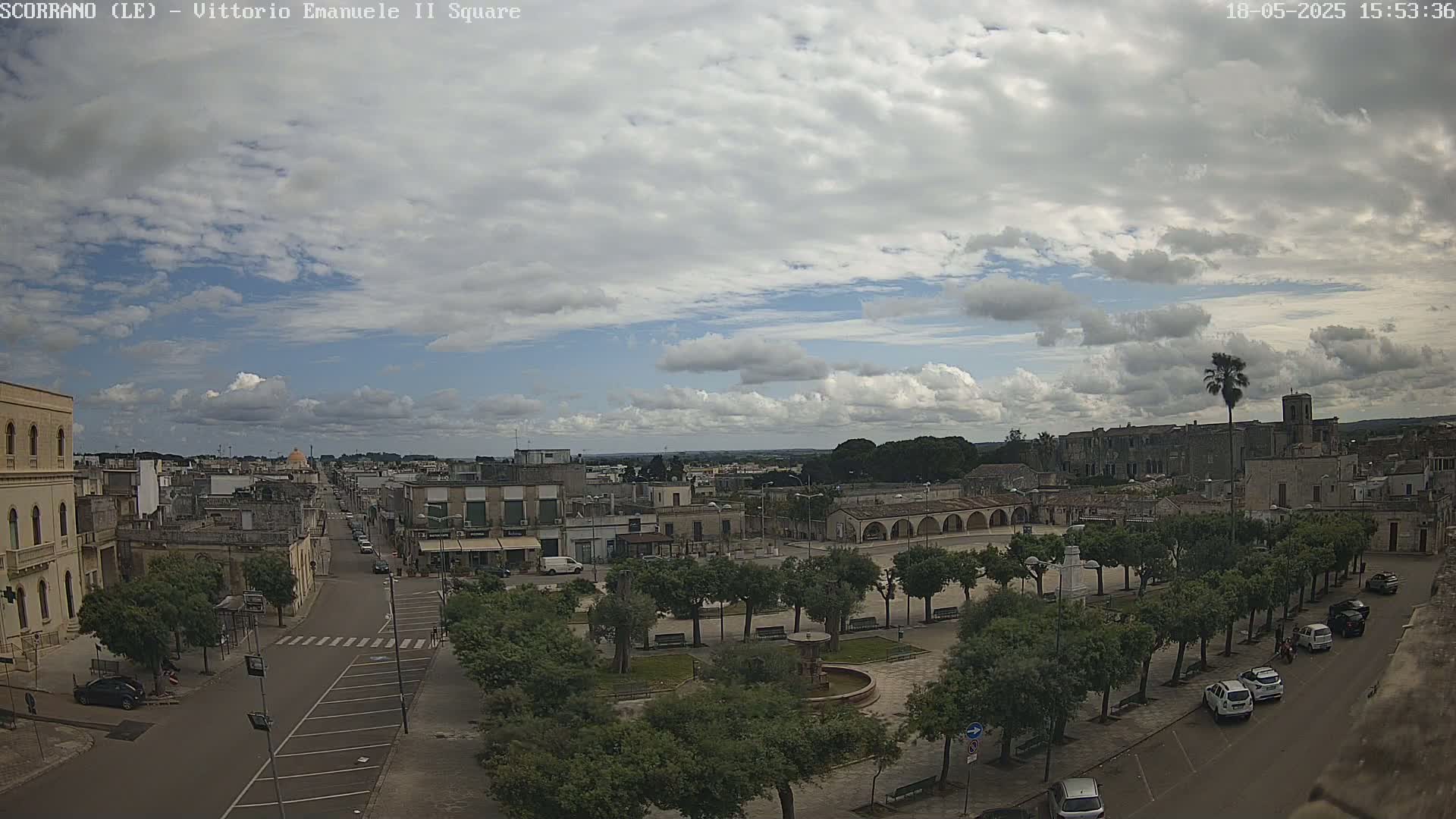 A partly cloudy sky overlooks a town square with a fountain and several parked cars, alongside a street with buildings and trees.