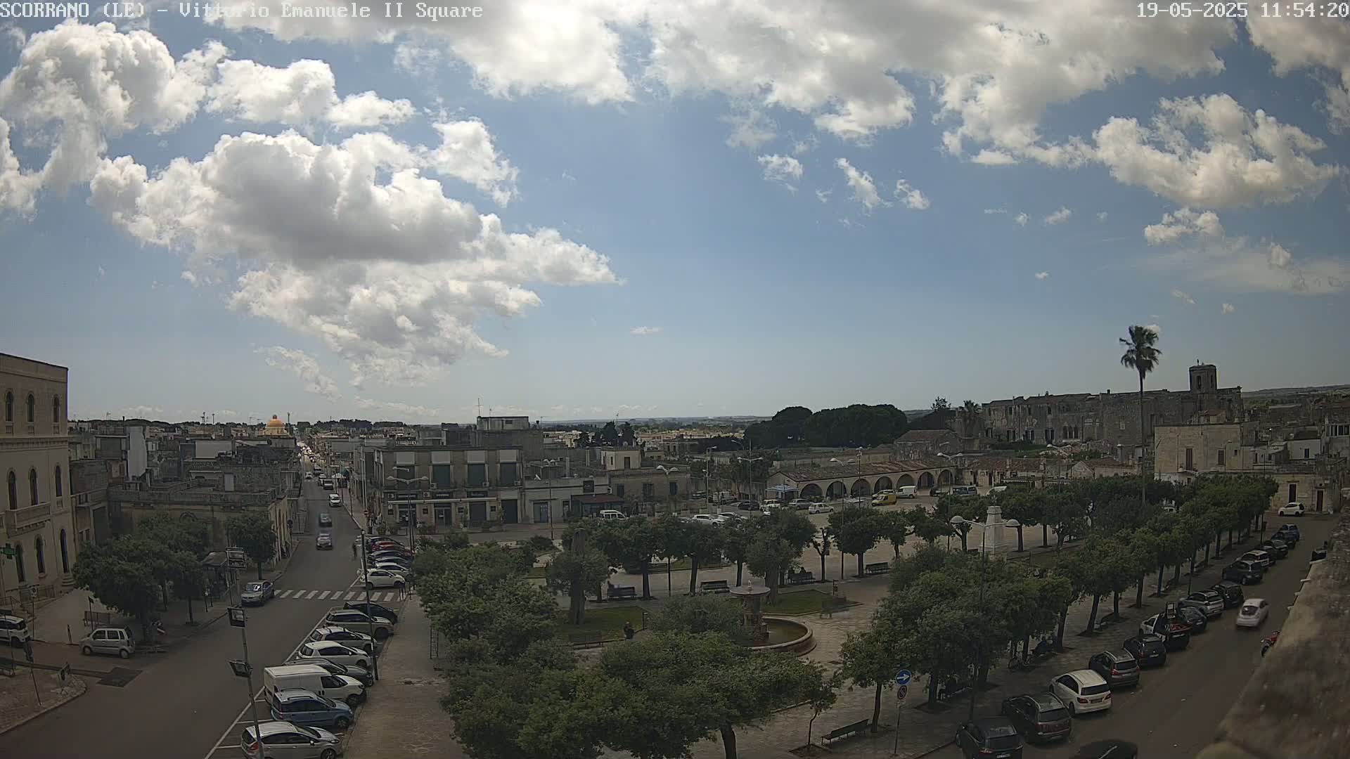 A partly sunny day reveals a town square with a fountain, surrounded by parked cars and trees, and streets lined with buildings extending into the distance.