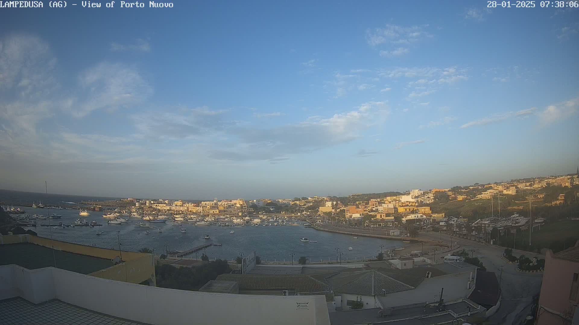 A harbor filled with boats is situated below a mostly clear, light blue sky with a few scattered clouds, overlooking a town built on a hillside.