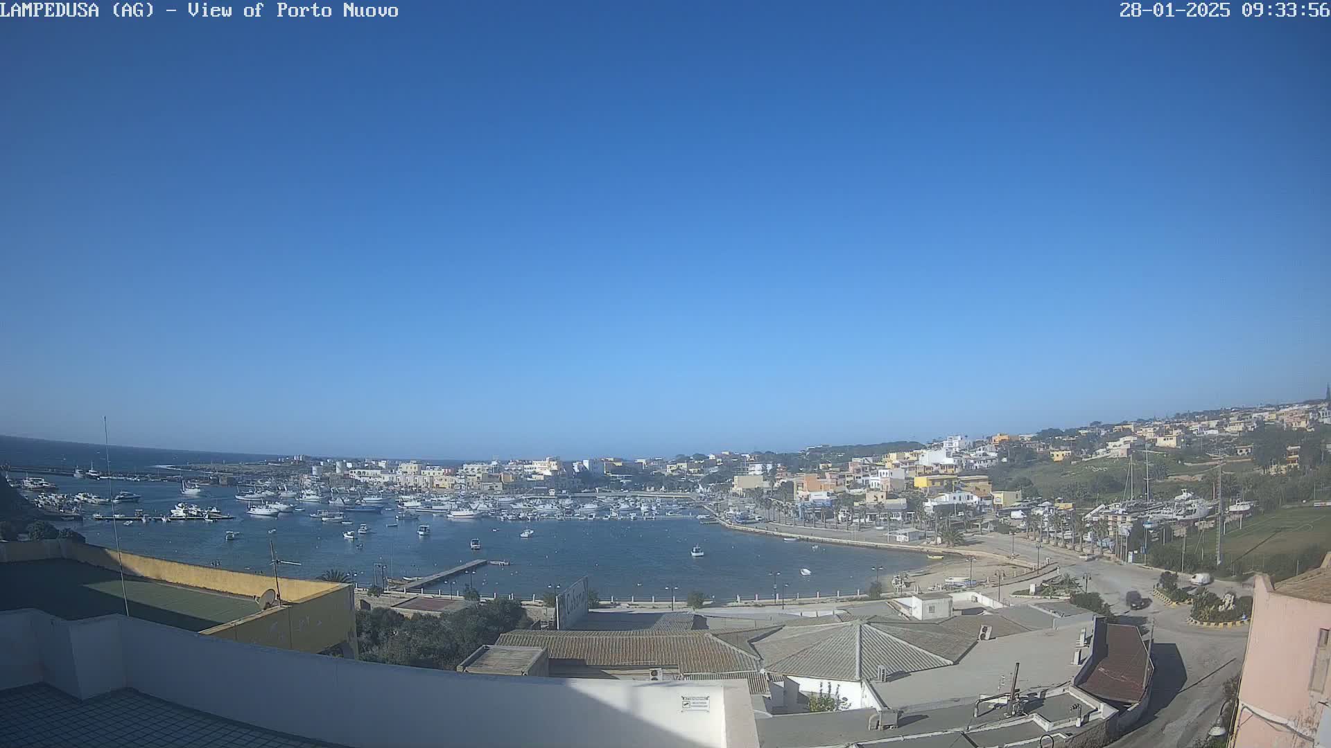 A harbor filled with boats is visible under a clear blue sky, with a town situated on a hillside in the background.