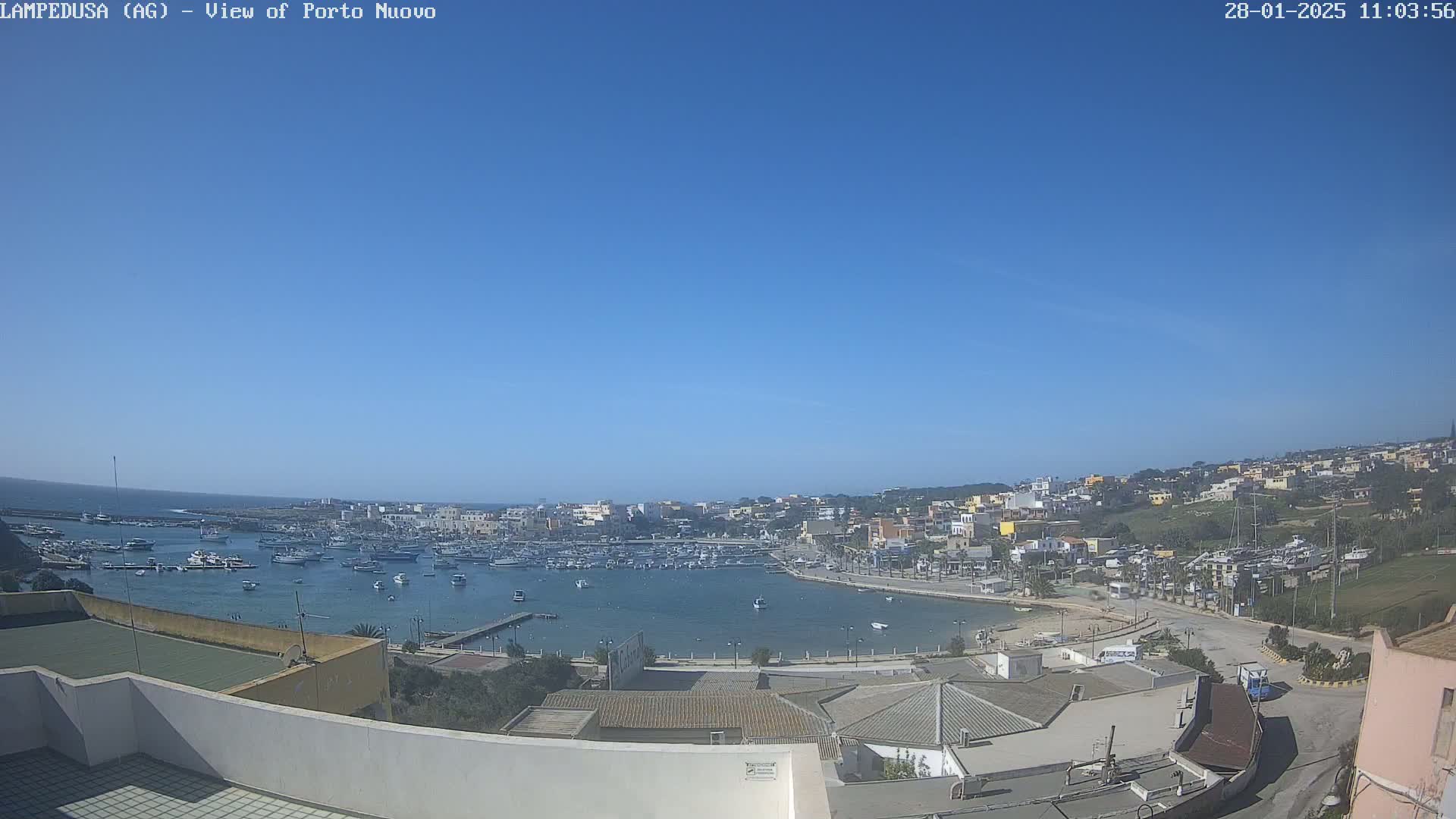A harbor filled with boats is situated in a town on a sunny day, with clear blue skies and hills visible in the background.