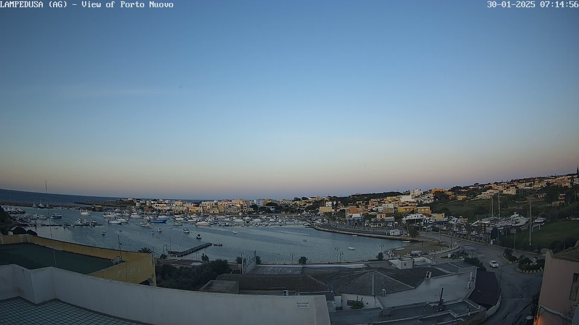 A calm harbor filled with boats is nestled within a town built on a hillside under a clear twilight sky.