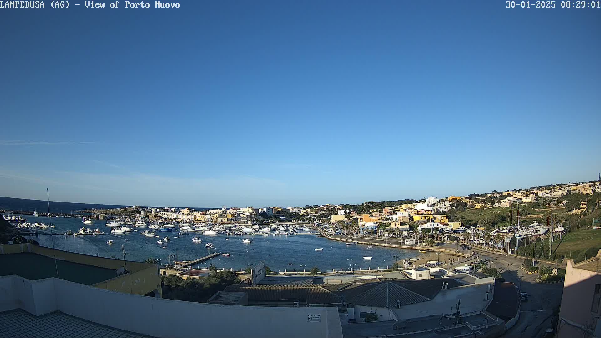 A harbor filled with numerous boats is nestled in a town of low-rise buildings under a clear blue sky.