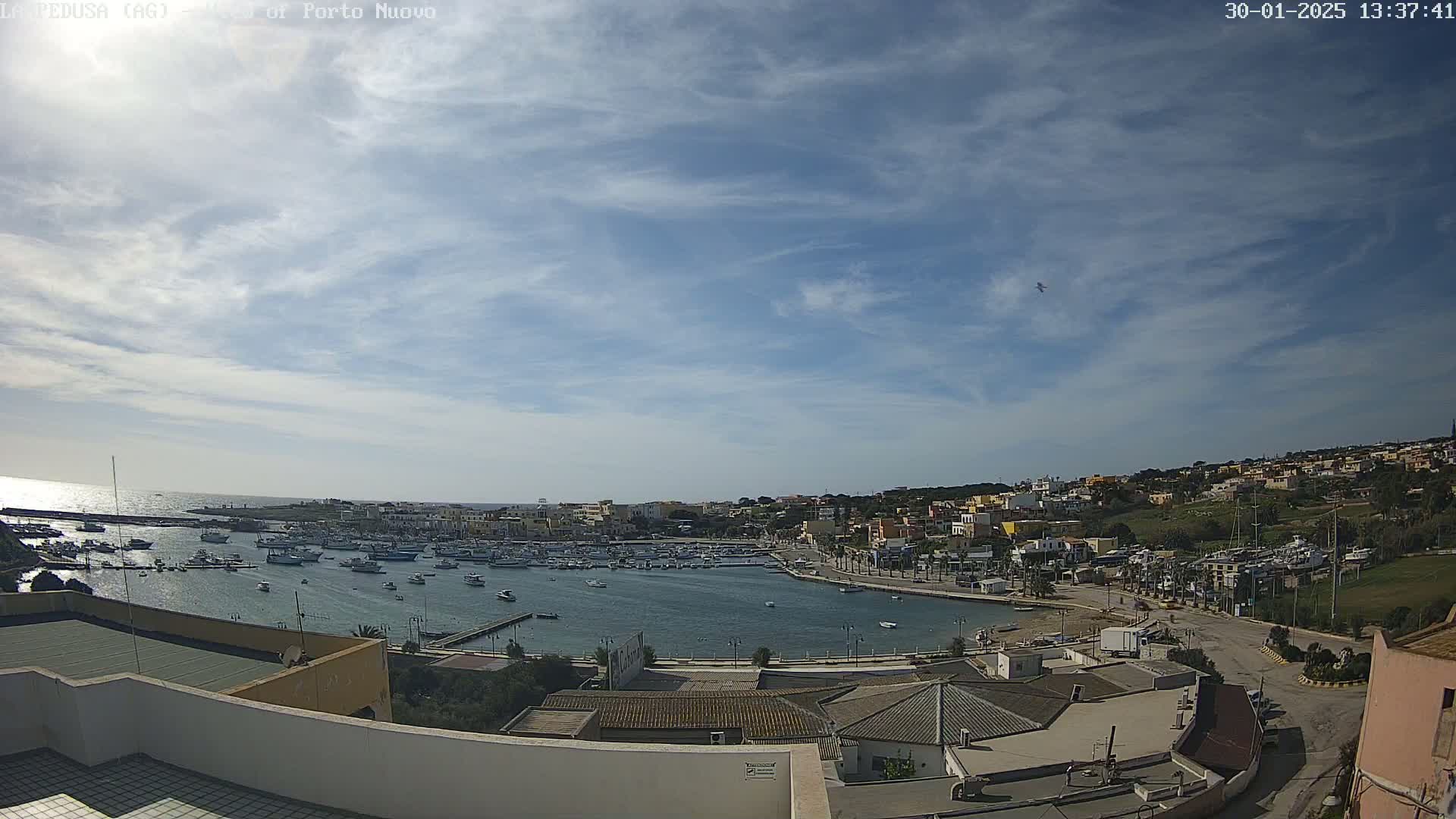 A harbor filled with boats is situated below a mostly clear sky with scattered clouds, overlooking a town built on a hillside.
