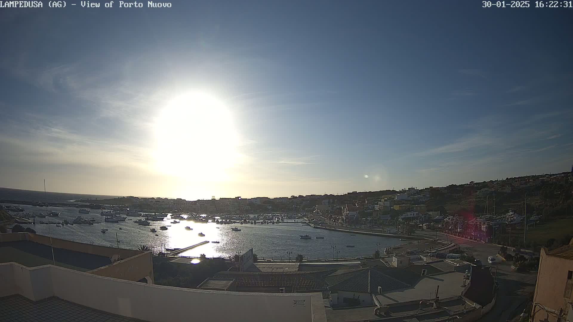 A sunny harbor scene shows numerous boats in calm water, with a town built on a hillside overlooking the water.