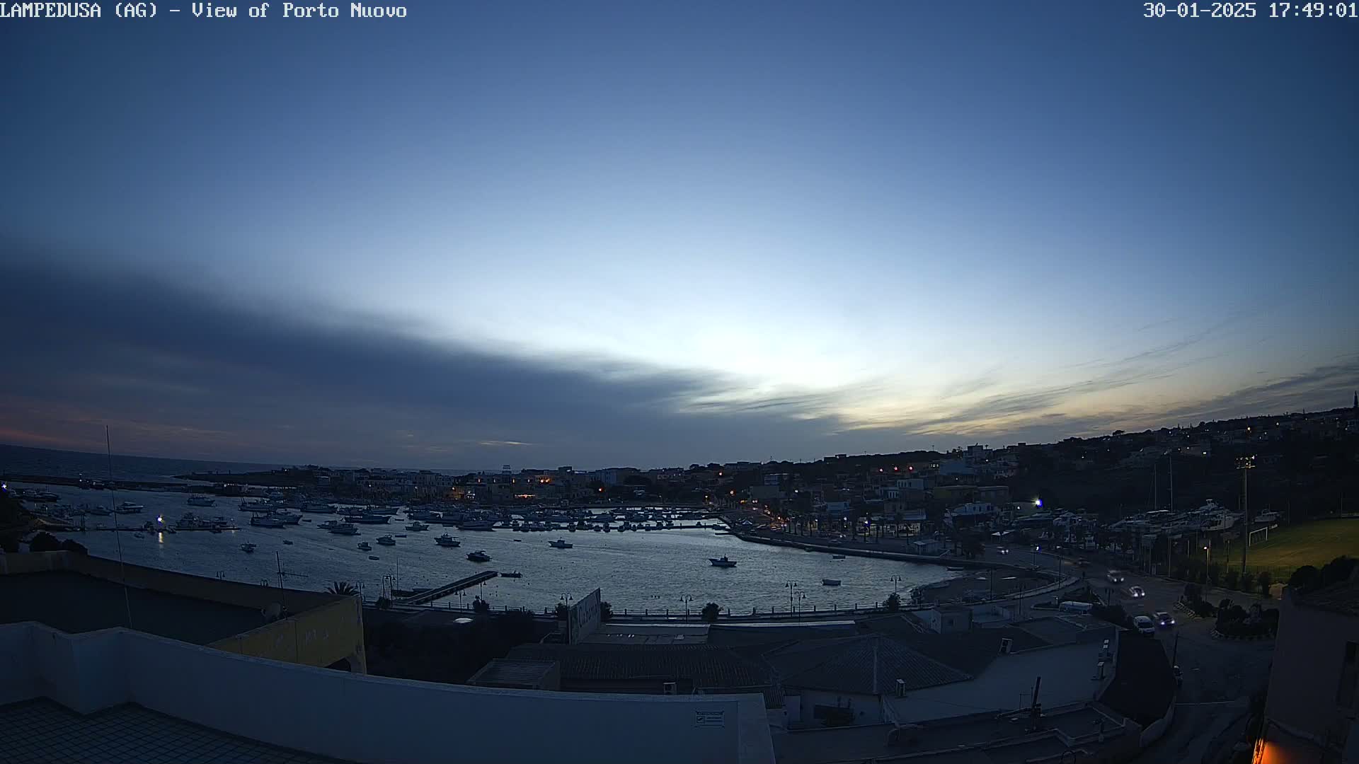 A harbor filled with boats is nestled in a town at twilight under a mostly clear, dusky sky.