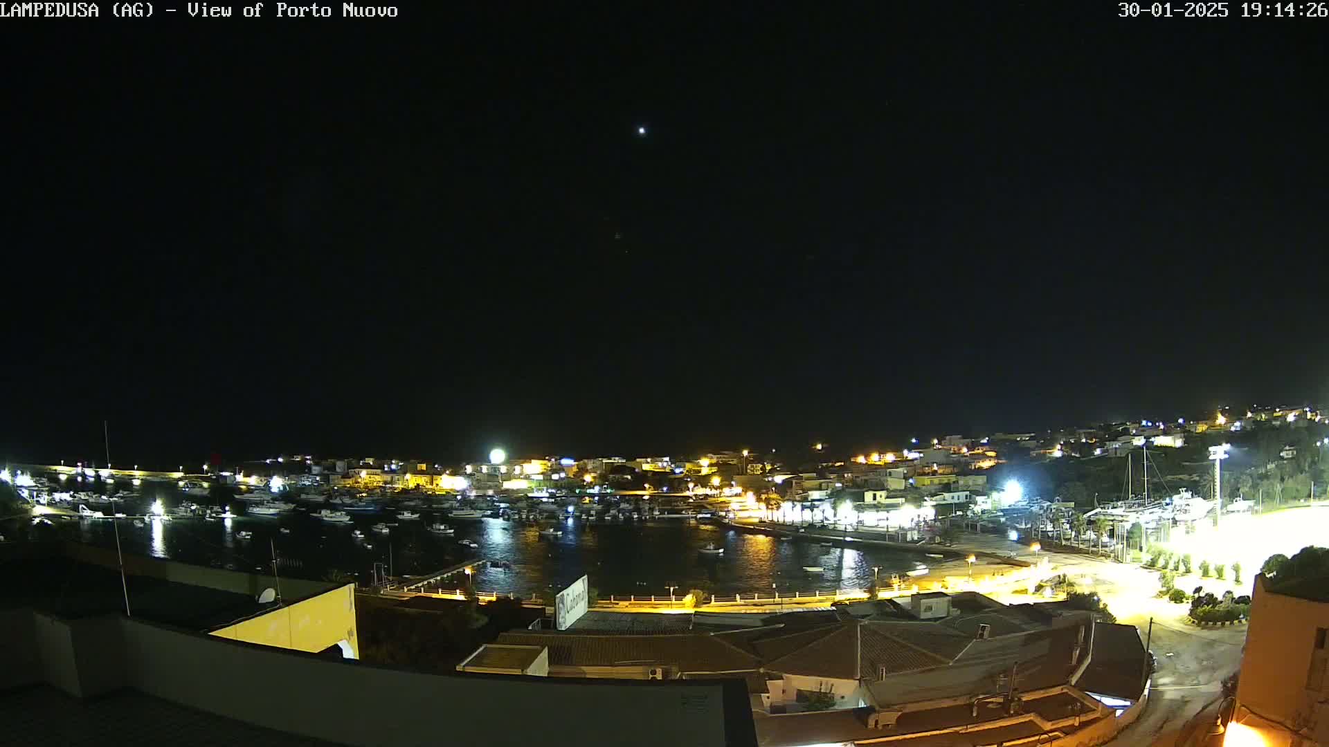A nighttime view of a harbor filled with boats, surrounded by buildings with lights illuminating the scene under a clear, dark sky.