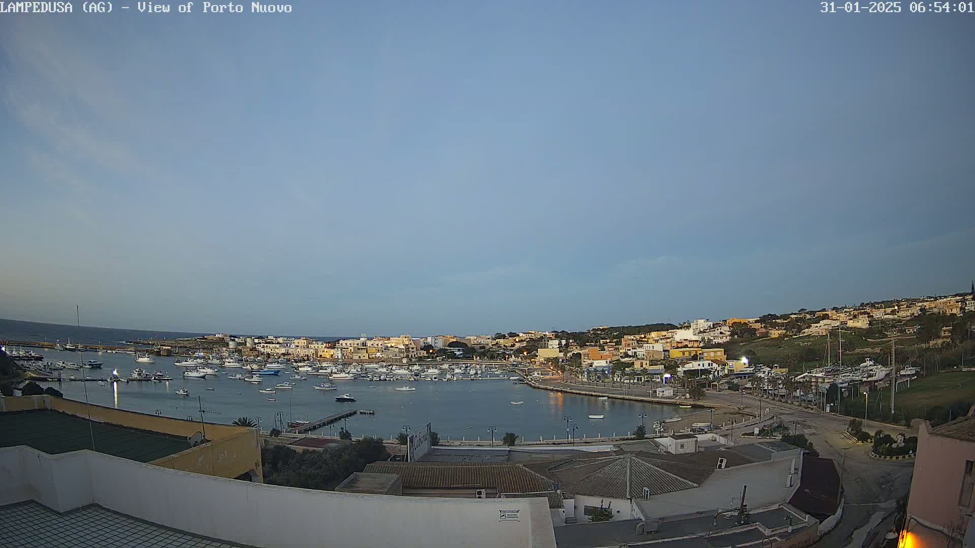 A harbor filled with boats is nestled within a town of low-lying buildings under a clear, pale blue sky.