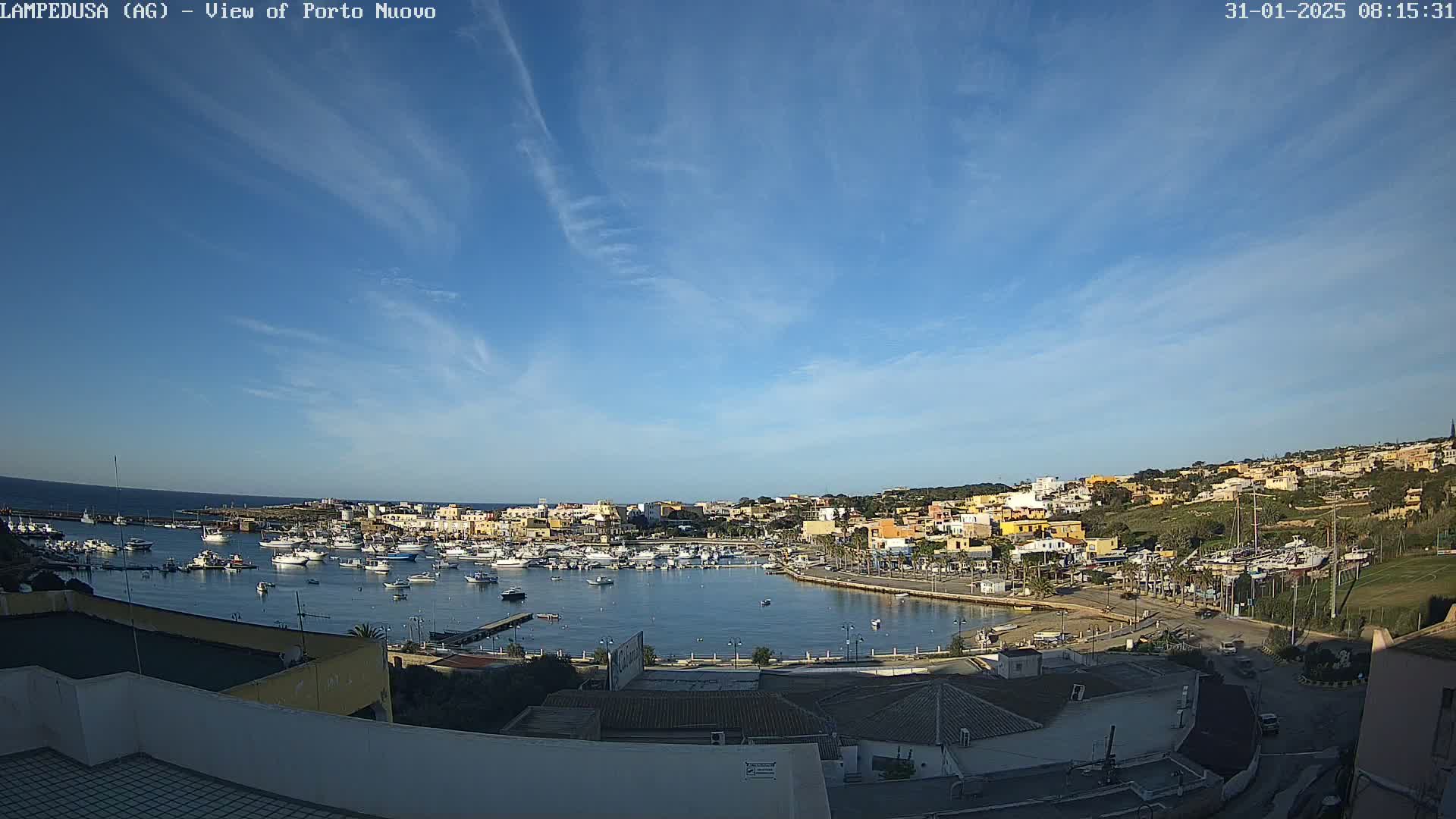 A harbor filled with boats is nestled in a town of low-rise buildings under a mostly clear, blue sky with wispy clouds.