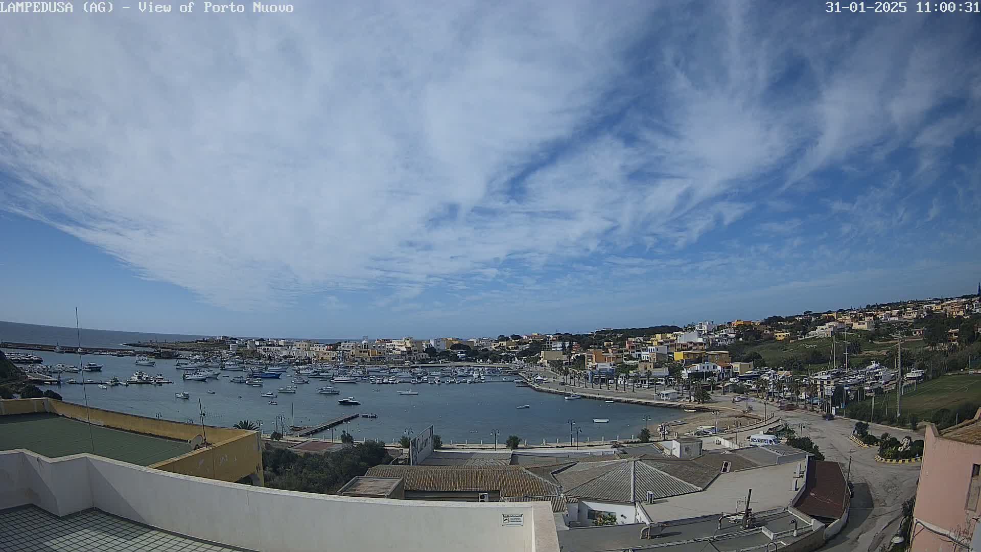 A harbor filled with boats is nestled in a town of low-rise buildings under a mostly sunny sky with scattered clouds.