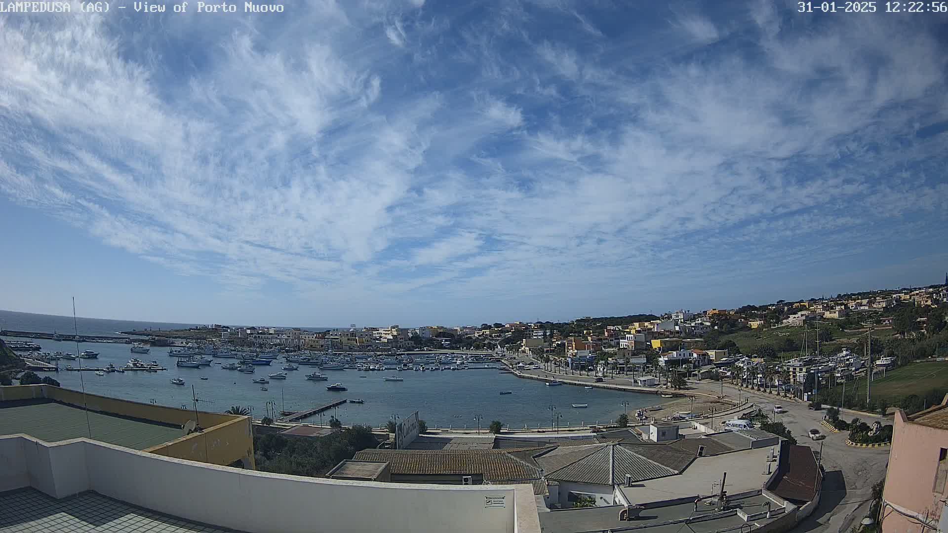 A harbor filled with boats is surrounded by a town under a mostly sunny sky with scattered cirrus clouds.