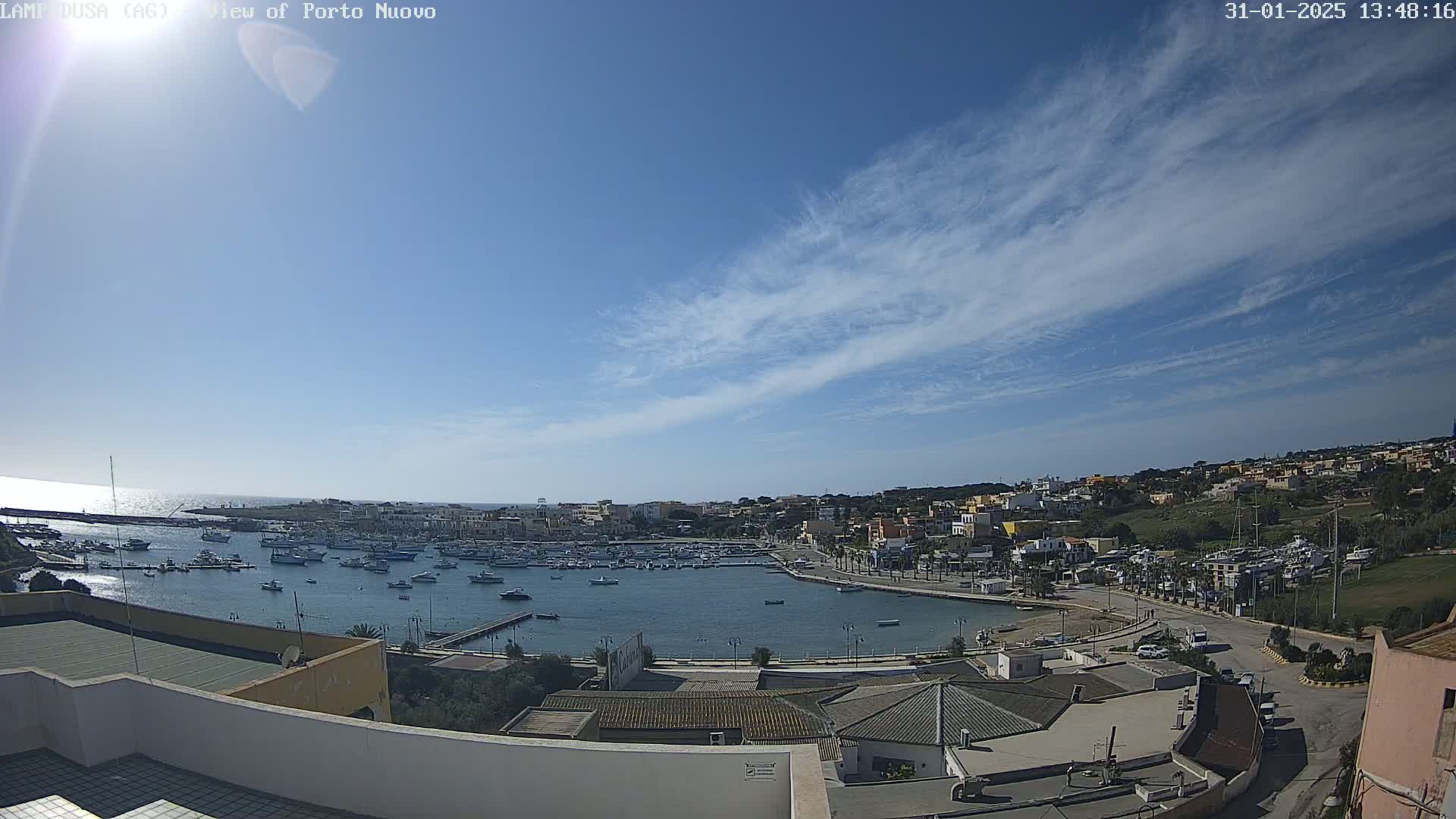 A harbor filled with boats is nestled in a town overlooking a calm sea under a mostly clear, bright sky with thin, wispy clouds.