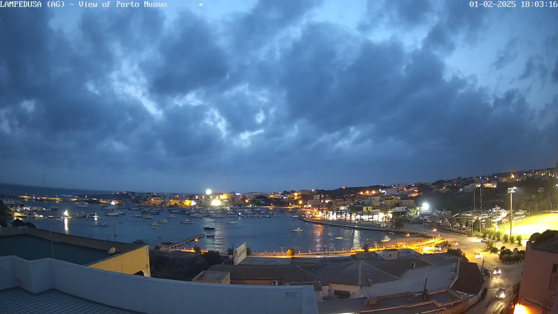 A nighttime harbor scene with a mostly cloudy sky shows a small town with illuminated buildings and boats in the water.