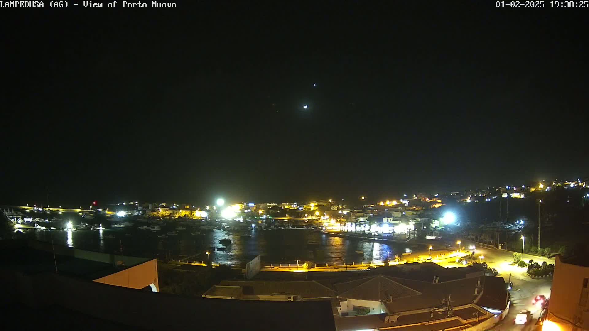 A nighttime, high-angle view of a harbor town with boats in the water and lights illuminating the buildings and streets under a clear, dark sky.
