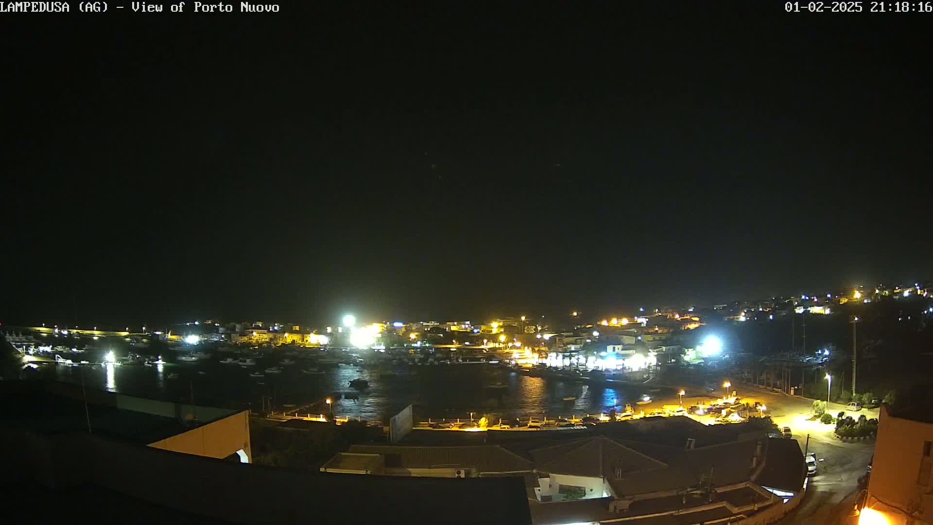 A nighttime aerial view shows a harbor filled with boats, surrounded by illuminated buildings and a dark, clear sky.