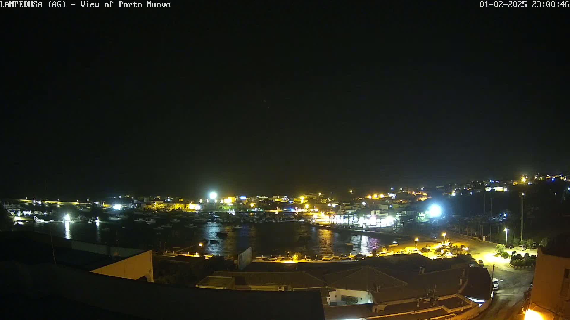 A nighttime, aerial view shows a harbor filled with boats and surrounded by illuminated buildings on a clear night.