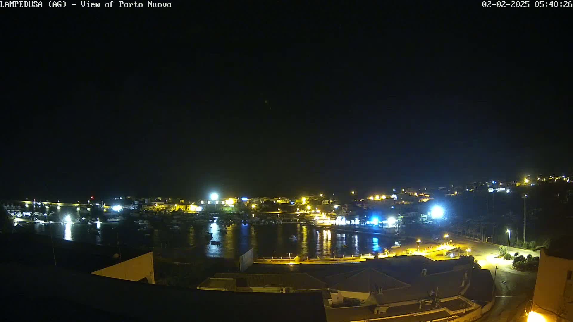 A nighttime, aerial view of a harbor filled with boats, illuminated by the lights of buildings along the shoreline under a clear, dark sky.