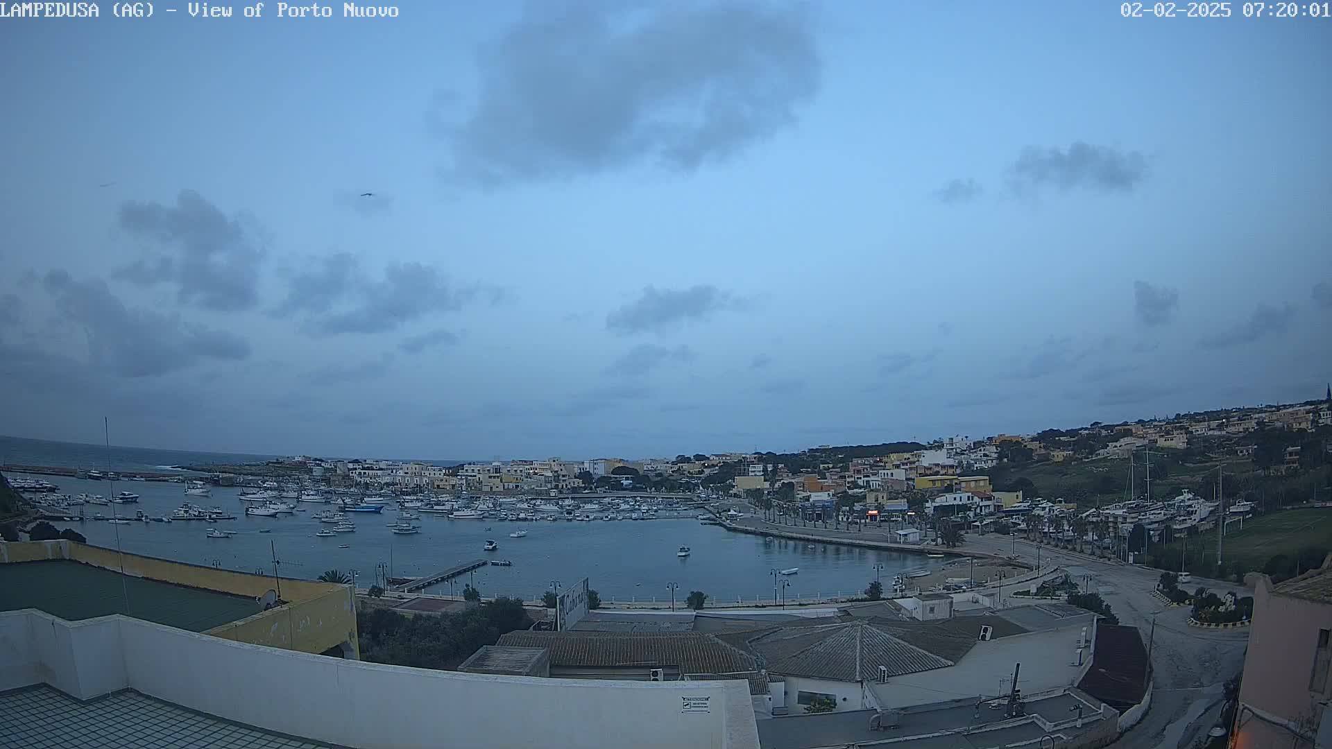 A harbor filled with boats is nestled in a town on a hill overlooking the ocean under a mostly cloudy, dusky sky.