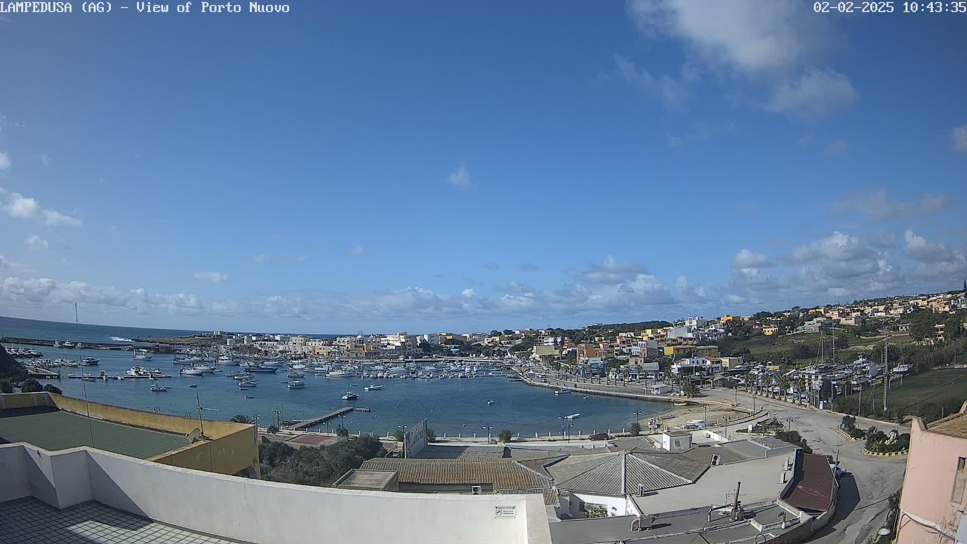 A harbor filled with boats is nestled in a town on a sunny day with partly cloudy skies.